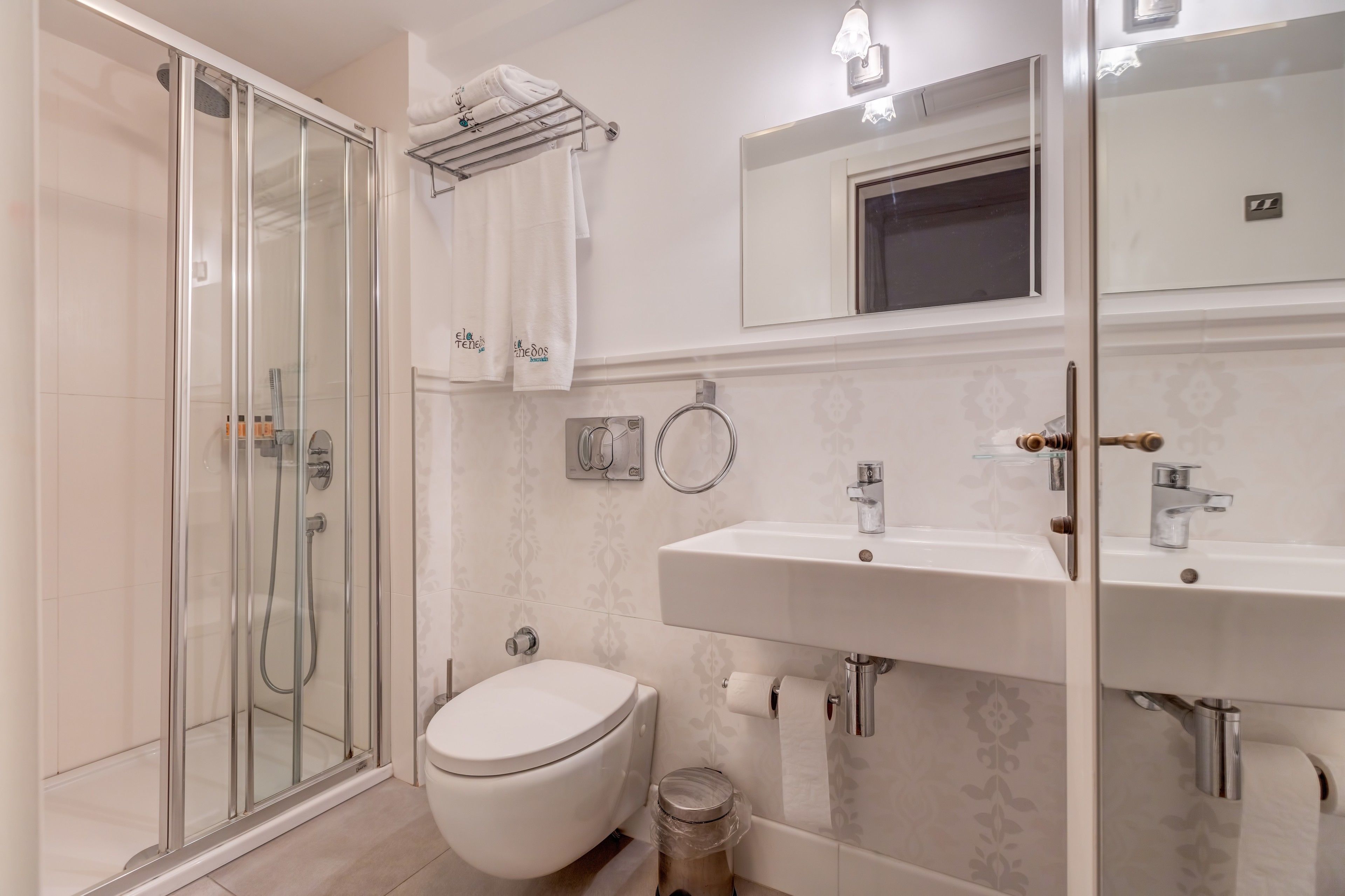 Un baño moderno con ducha envuelta en vidrio, inodoro blanco, lavabo con espejo y perchero con toallas.