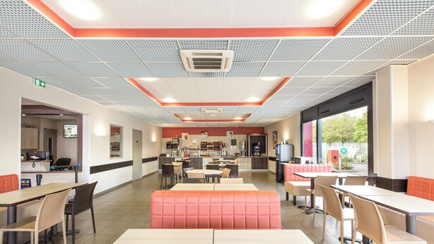 A modern restaurant with a central seating area, a kitchen in the background, and a large window providing natural light. - ACE Hôtel Chartres (Le Coudray)
