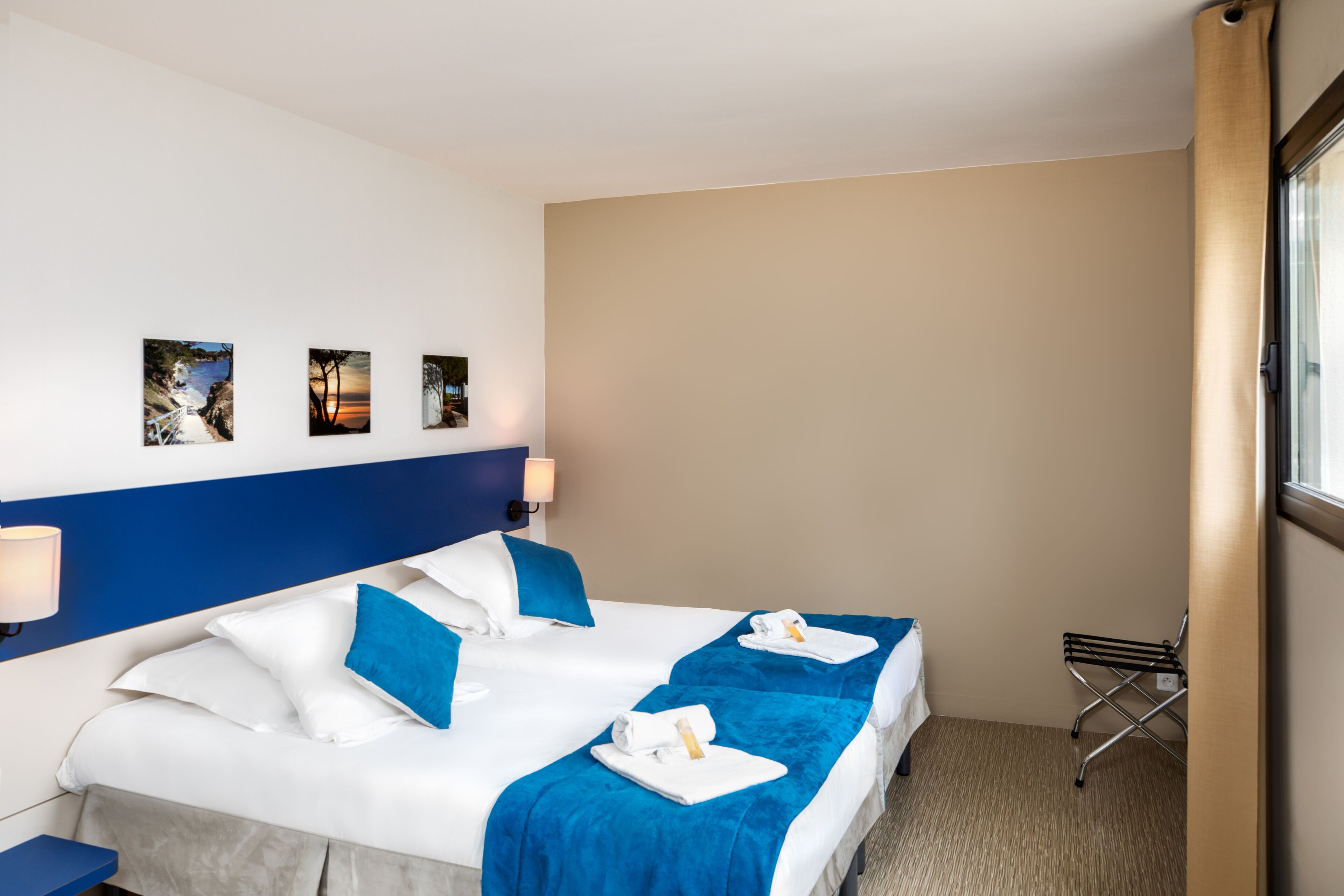 A hotel room with a large bed, two bedside tables, a window with curtains, and three wall-mounted pictures.