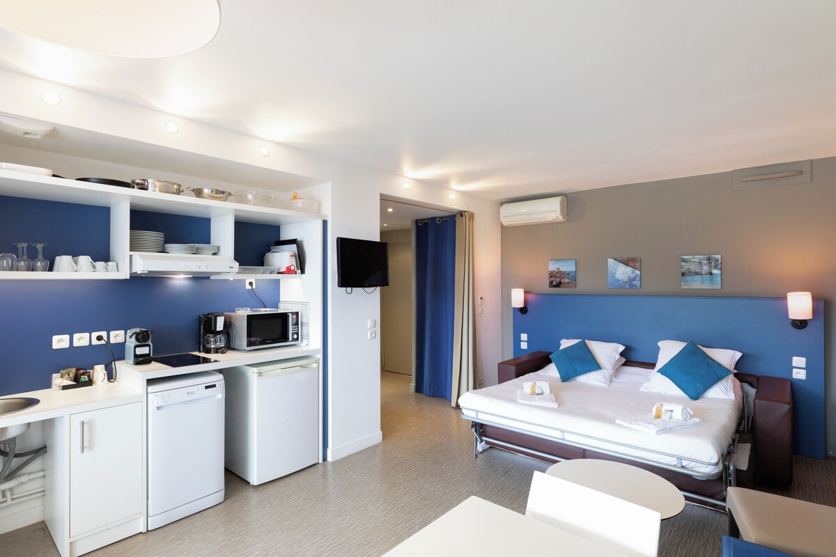 Une chambre d’hôtel compacte comprenant un coin cuisine, un lit avec des oreillers bleus, une table de chevet et un téléviseur.