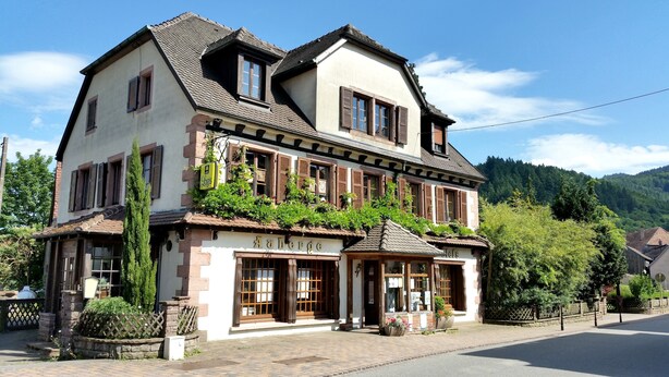 Un bâtiment à deux étages de style architectural européen traditionnel, doté d’un balcon orné de plantes, d’une enseigne indiquant un restaurant et d’une entrée bien visible avec une porte en bois.