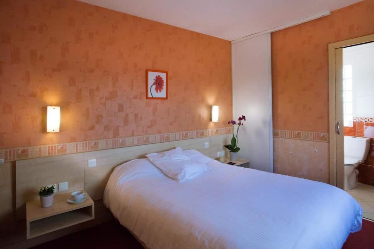 Habitación de hotel con cama, mesitas de noche, lámpara de pared, un cuadro enmarcado y una planta en maceta.