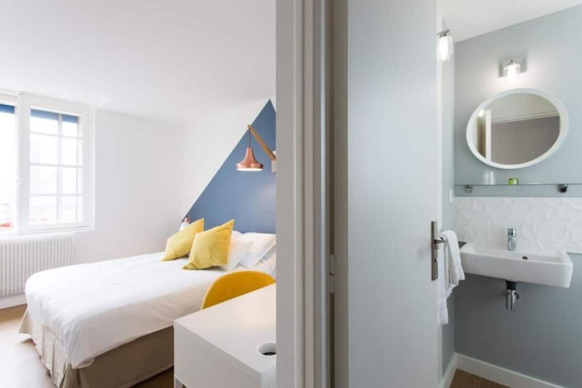 Un dormitorio con cama, mesita de noche, ventana y baño con lavabo y espejo.