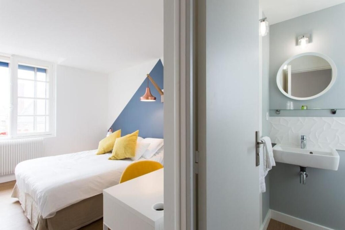 A bedroom with a bed, a bedside table, a window, a bathroom with a sink and mirror.