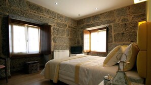 Une chambre avec des murs en pierre, un lit avec une literie blanche, une table de chevet en bois, une télévision et une fenêtre avec des rideaux.