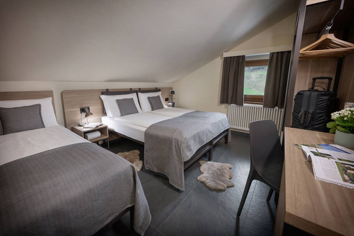 A hotel room with two beds, a desk, a chair, a radiator, and a window with curtains.