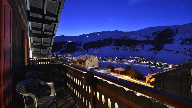 A balcony with a view of a snow-covered mountain and a lit town below. - Hotel Loredana (Livigno)