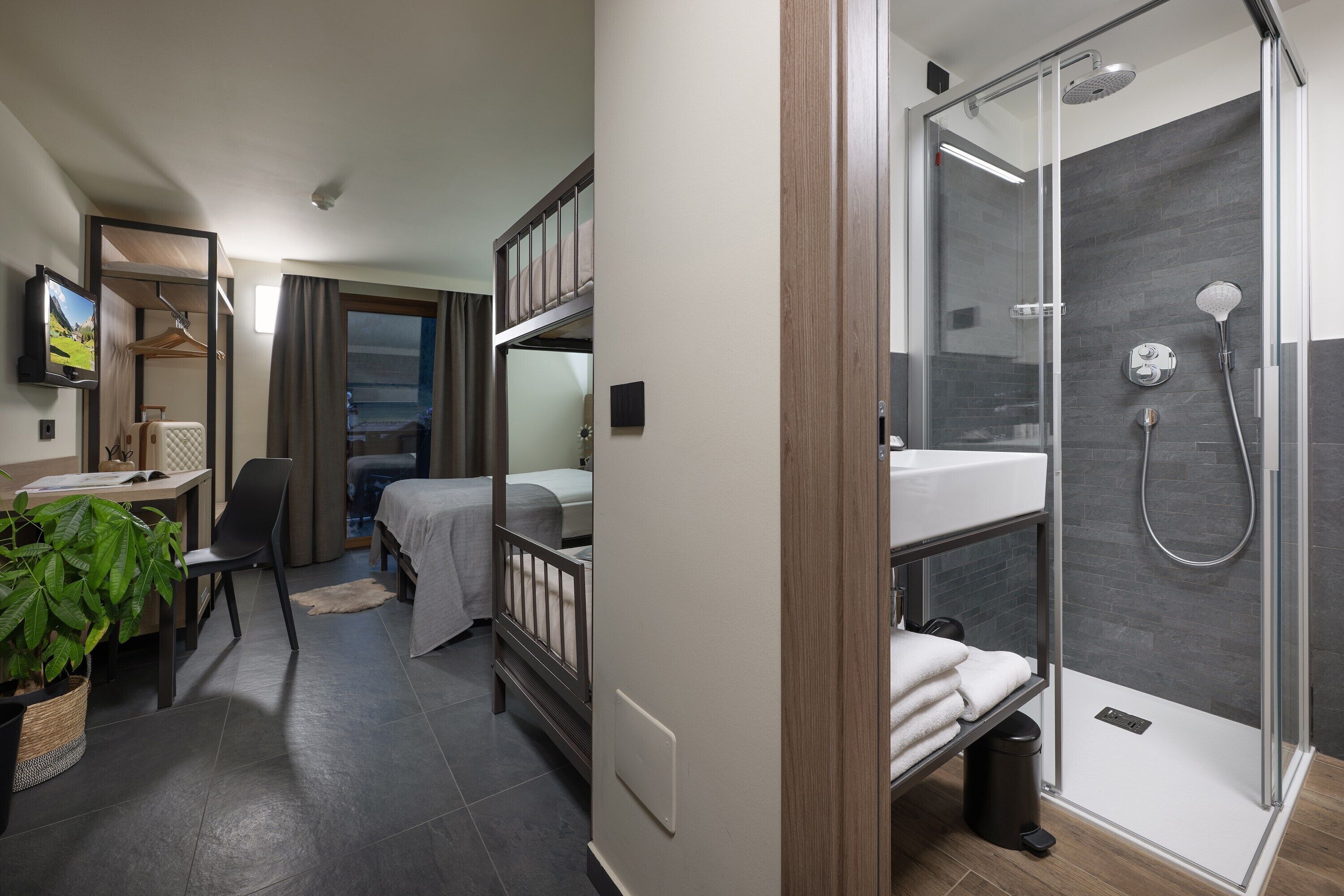 A hotel room with a bed, a desk, a chair, a potted plant, a shower with a glass door, and a view of the outdoors.