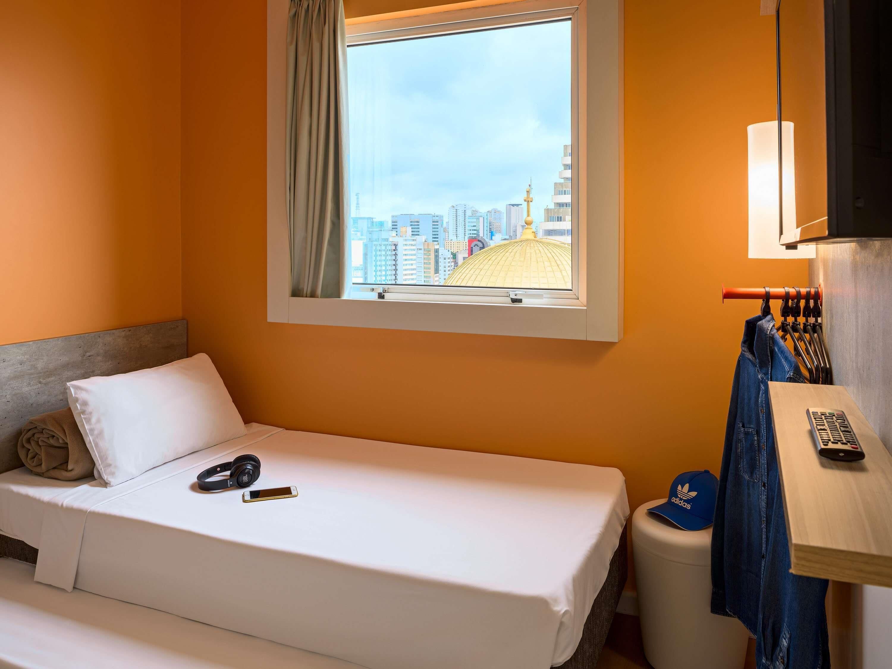 Quarto de hotel com cama, janela com vista para a cidade, escrivaninha com abajur e uma cadeira.