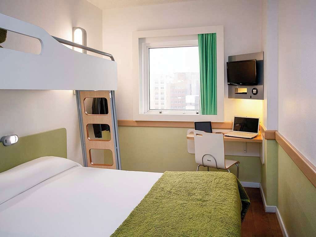 A hotel room with bunk beds, a desk with a computer, a TV, and a window with a curtain.