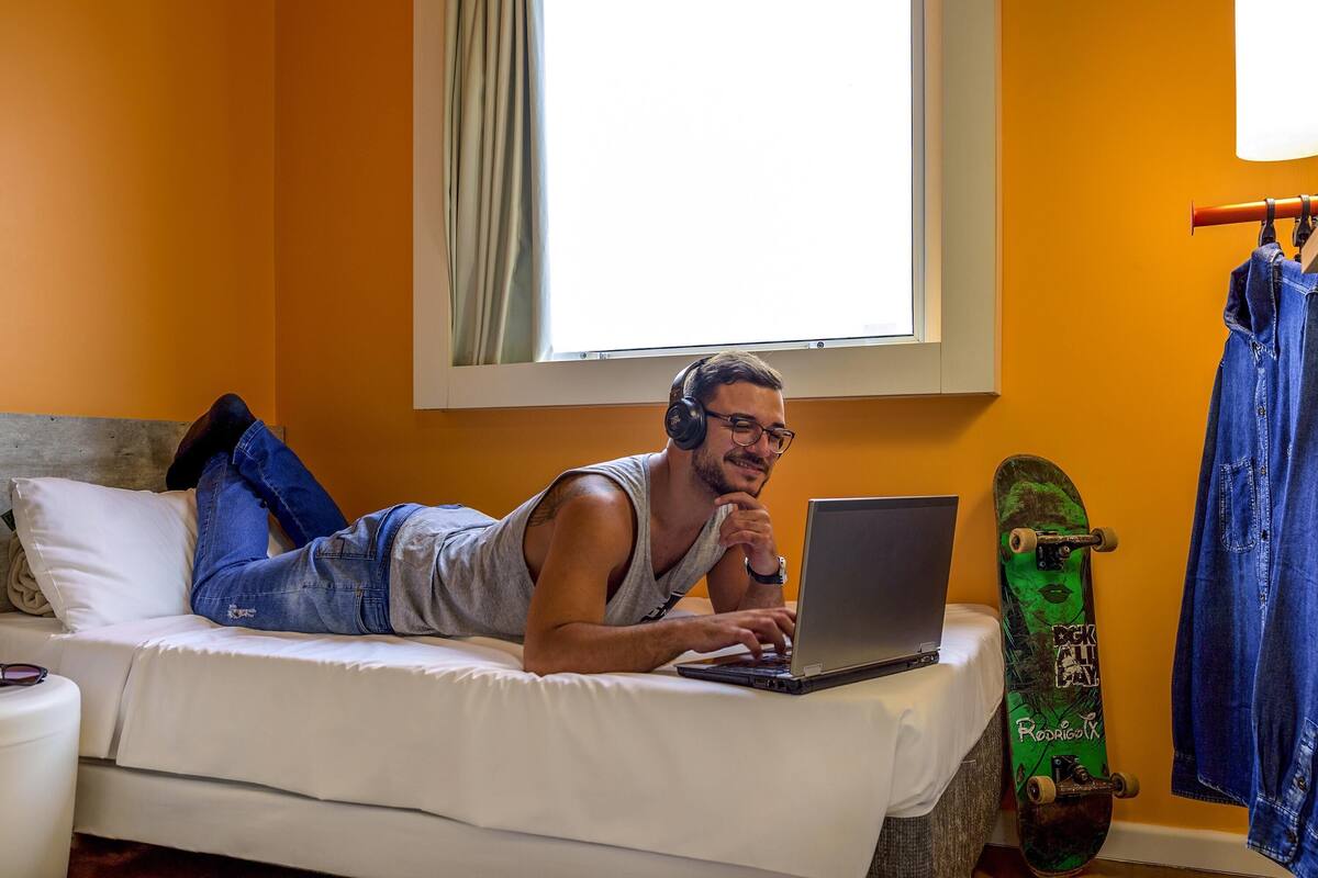Een persoon ligt op een bed en gebruikt een laptop, met een skateboard en een spijkerjasje in de buurt.