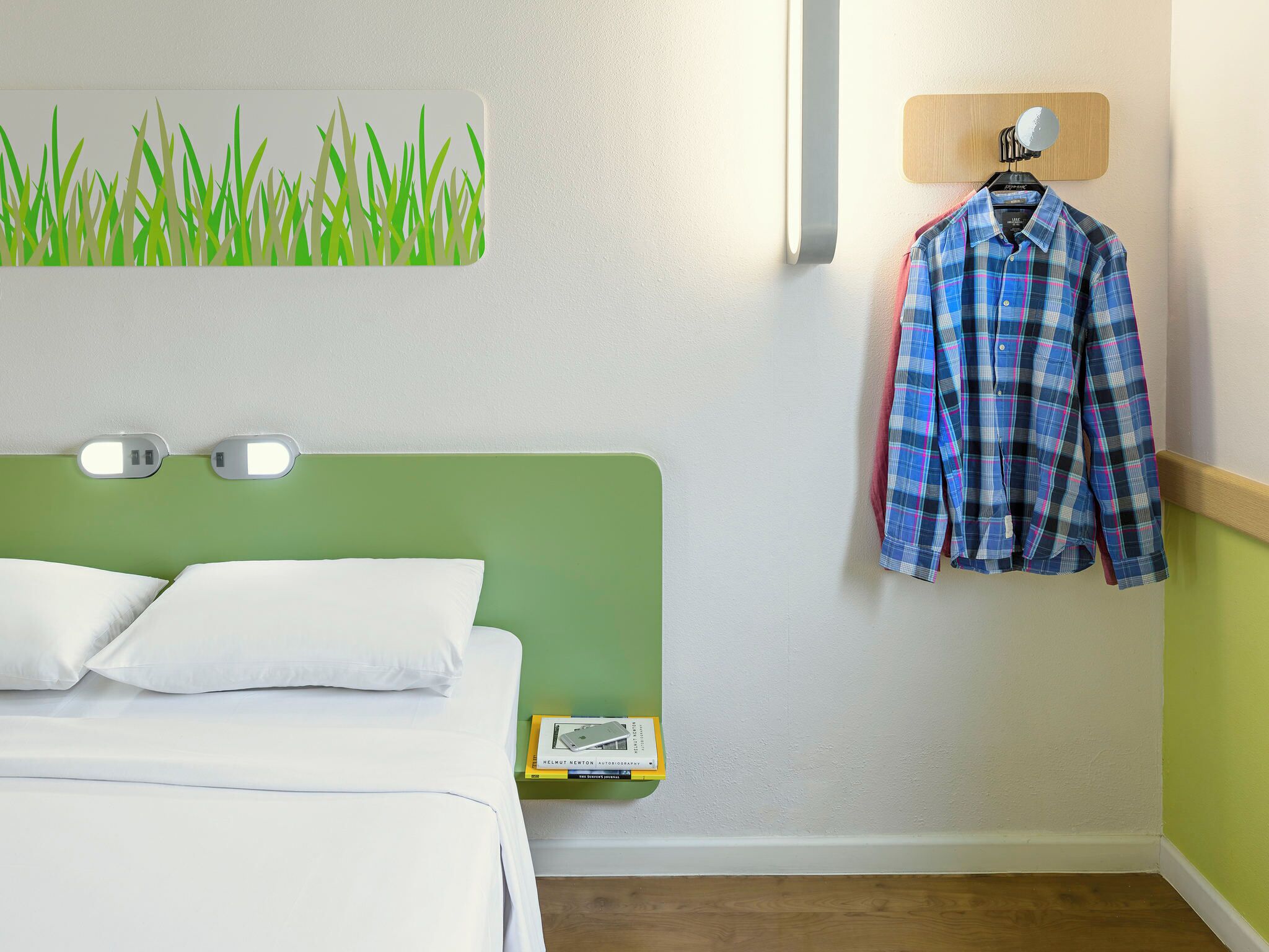 Habitación de hotel con cama, almohadas, cabecera verde, espejo montado en la pared, camisa escocesa colgada y una pequeña estantería con libros y un teléfono.