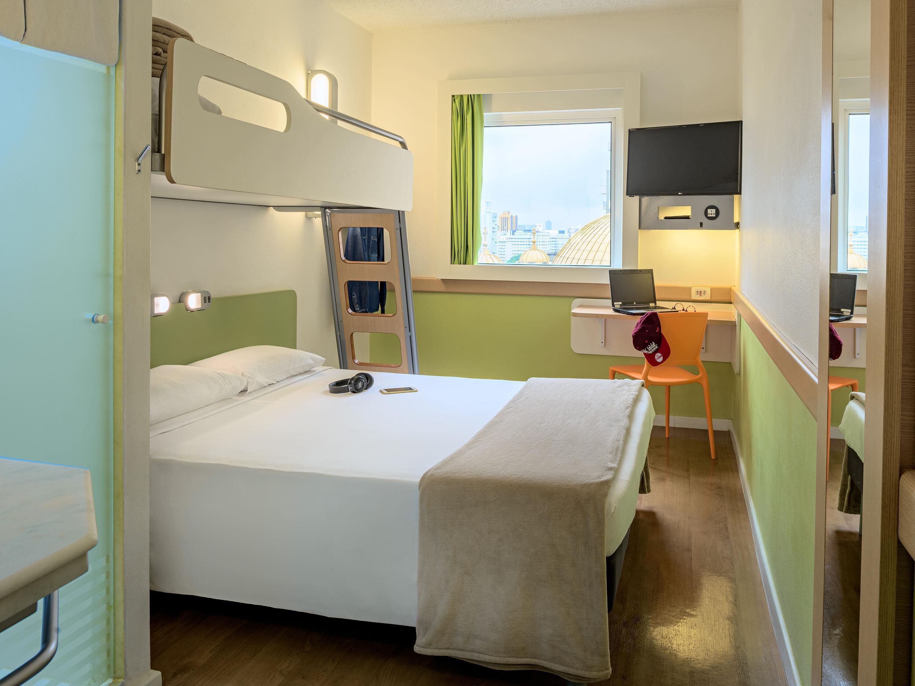 Habitación de hotel con literas, un escritorio con una computadora portátil, una silla, una ventana con vistas a edificios y una cama con una manta.