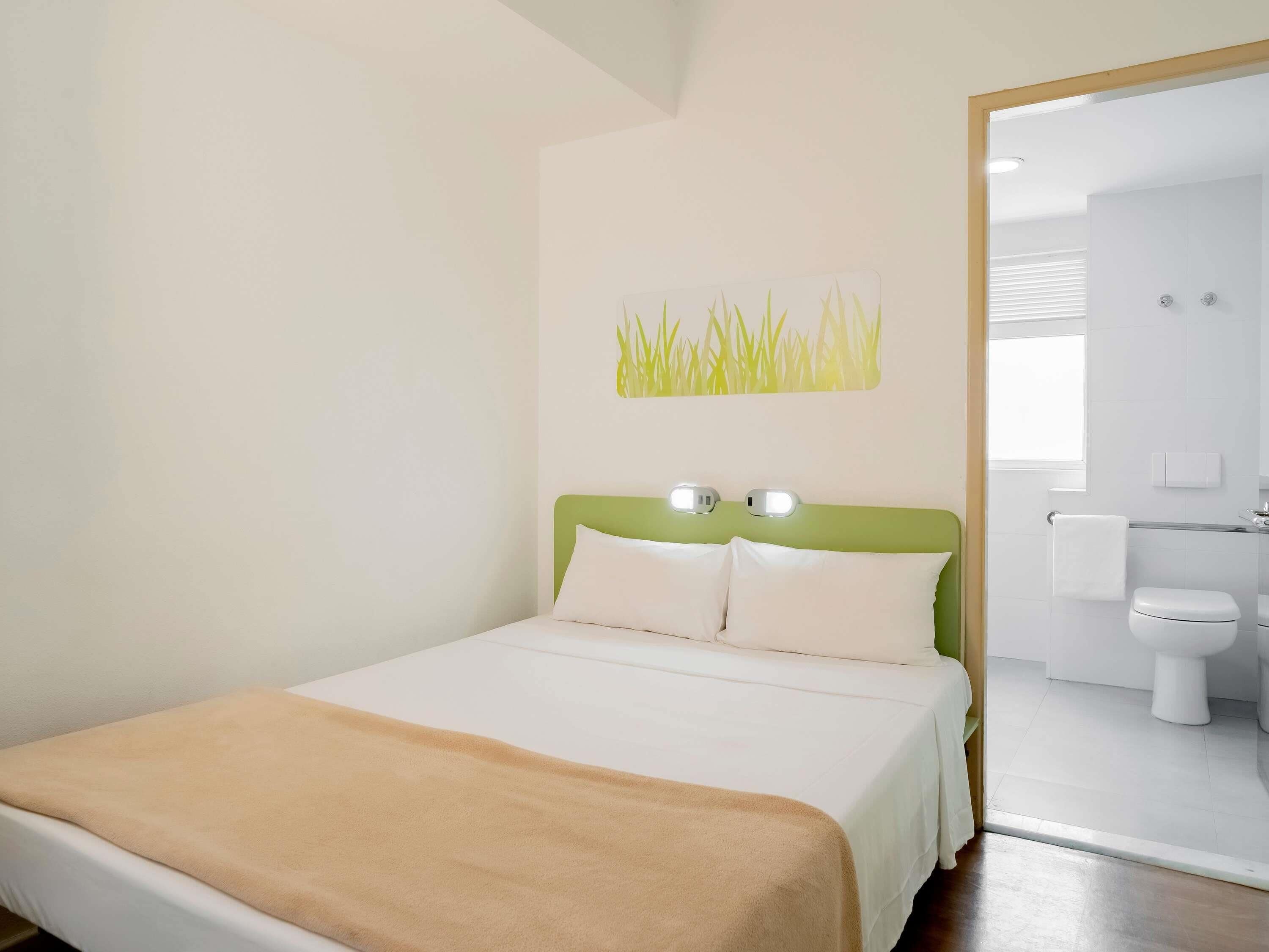 A hotel room with a bed, a green headboard, two bedside lamps, a white bathroom with a toilet and sink, and a window.
