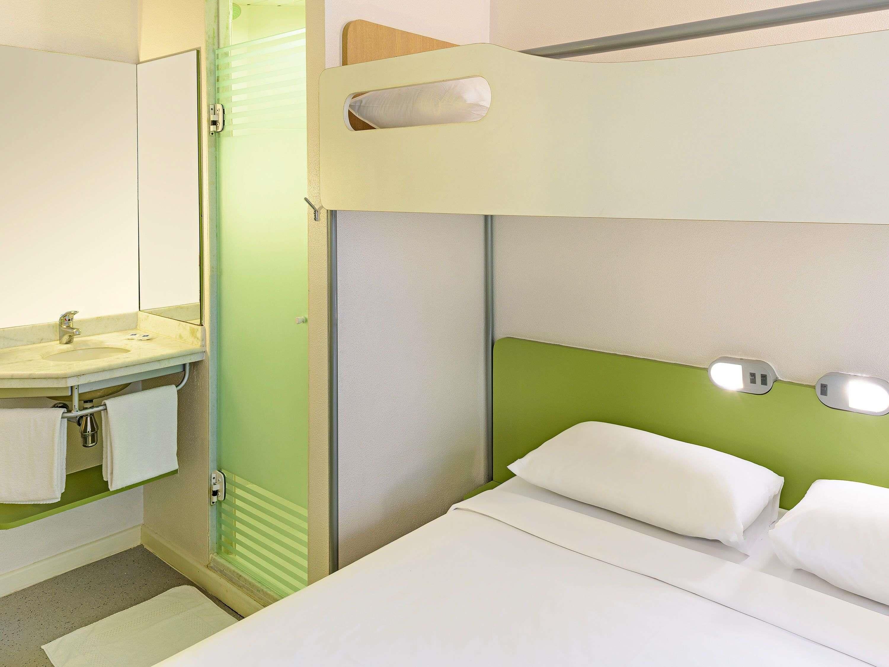 A hotel room with a bunk bed, a bathroom with a sink and mirror, and a bedside table with a lamp.