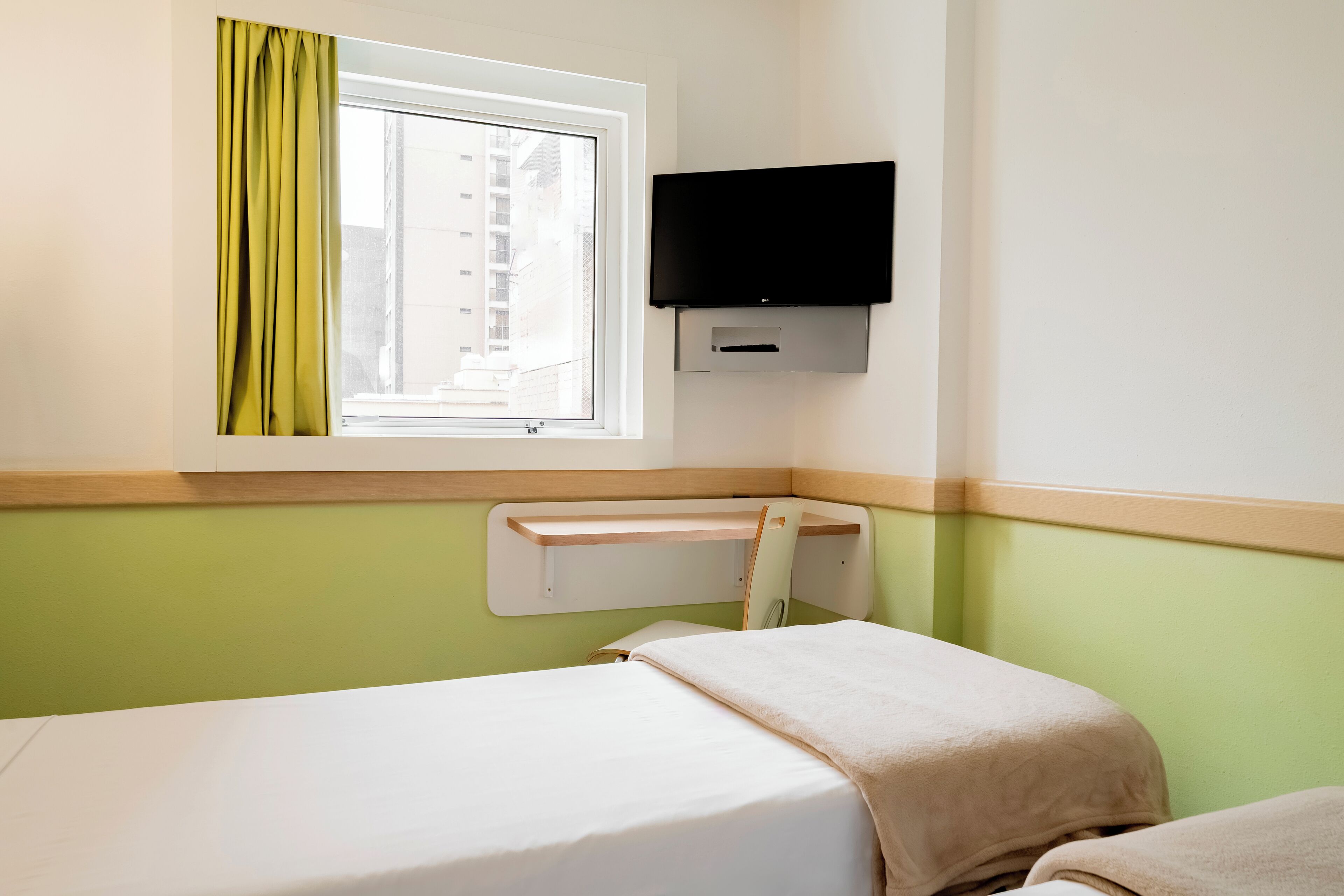 A hotel room with a bed, a window with curtains, a mounted TV, and a small shelf.