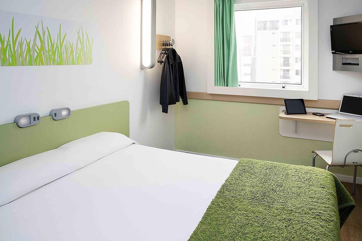 Une chambre d’hôtel avec un lit, un bureau avec un ordinateur portable, une télévision et une fenêtre avec des rideaux.