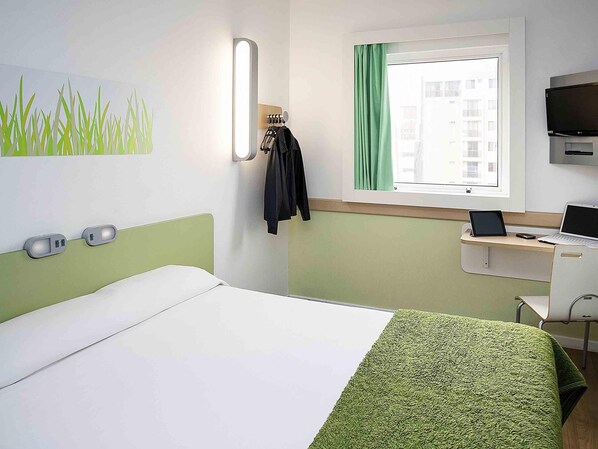 A hotel room with a bed, a desk with a laptop, a TV, and a window with curtains.