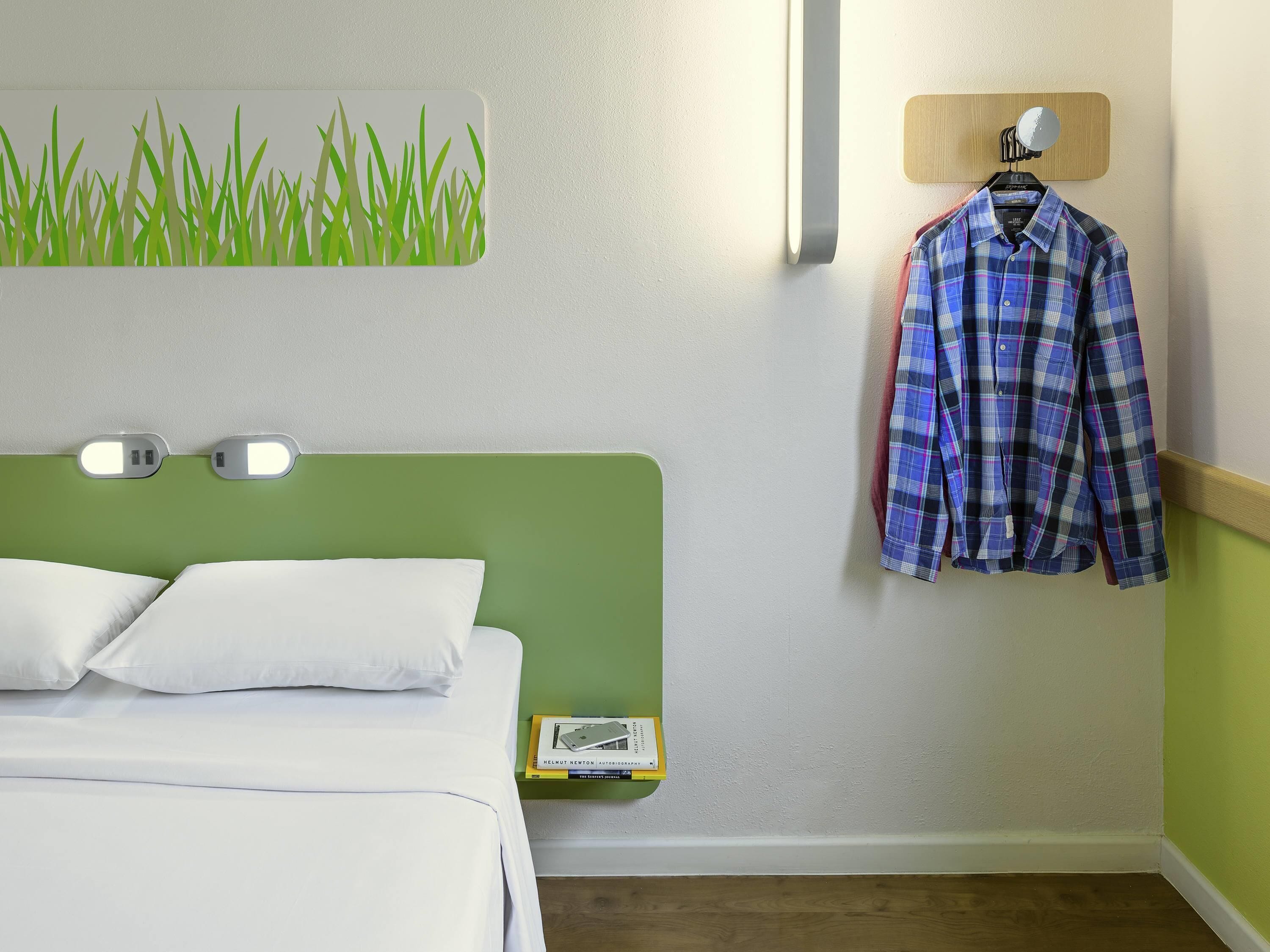 Quarto de hotel com cabeceira verde, roupa de cama branca, luminária fixada na parede, camisa pendurada e uma pequena mesa de cabeceira com um livro.