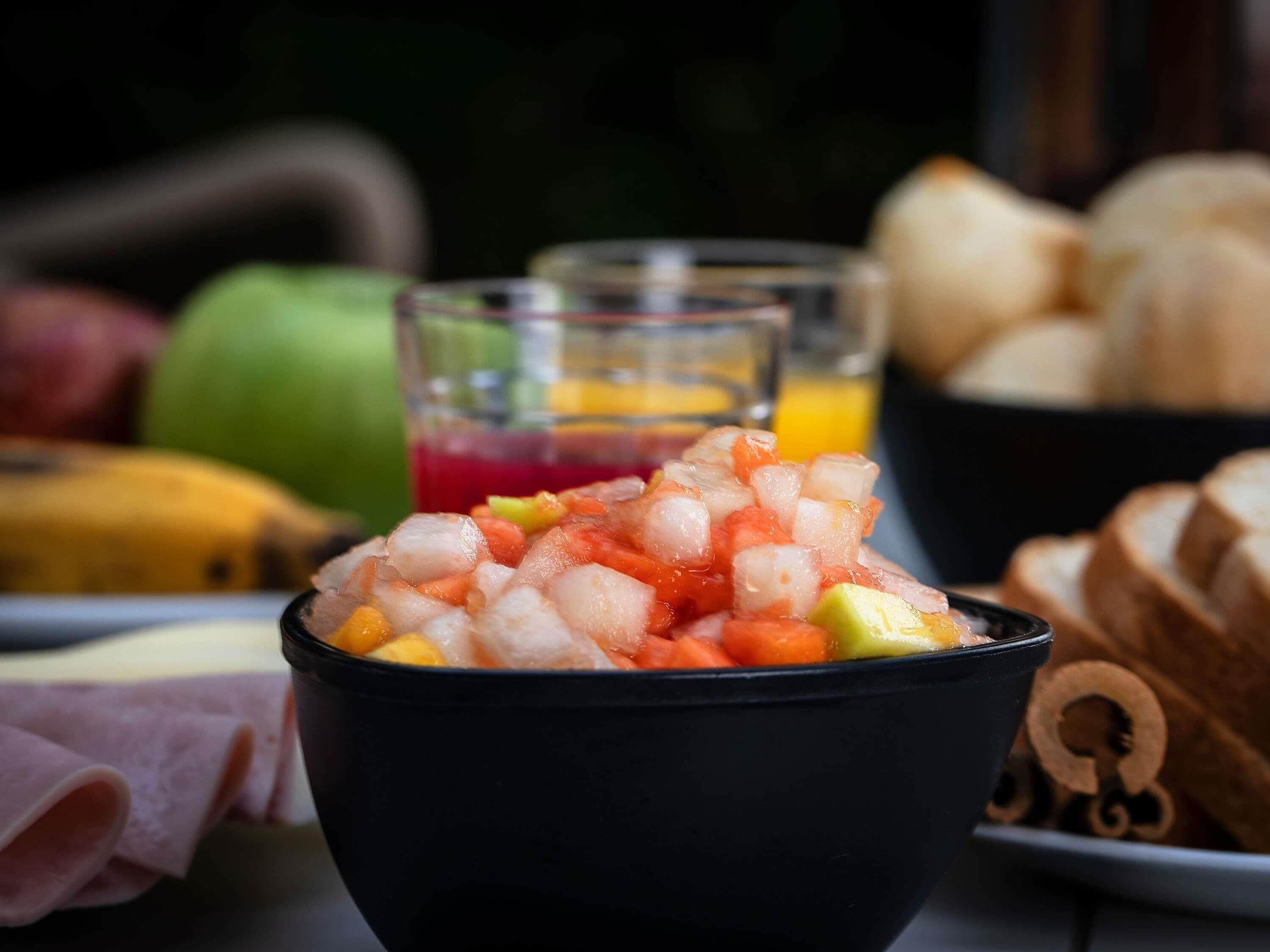 A bowl of diced fruit and ice, a glass of red juice, and bread on a table.