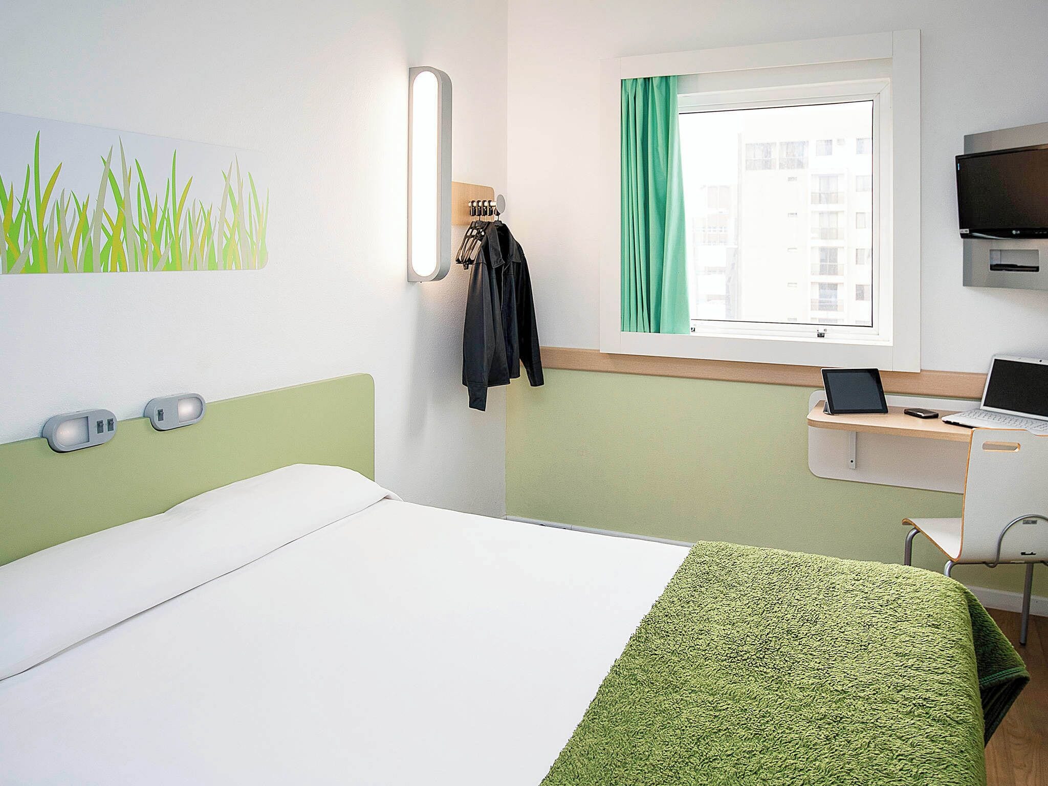 A hotel room with a bed, a desk with a laptop, a TV, and a window with curtains.