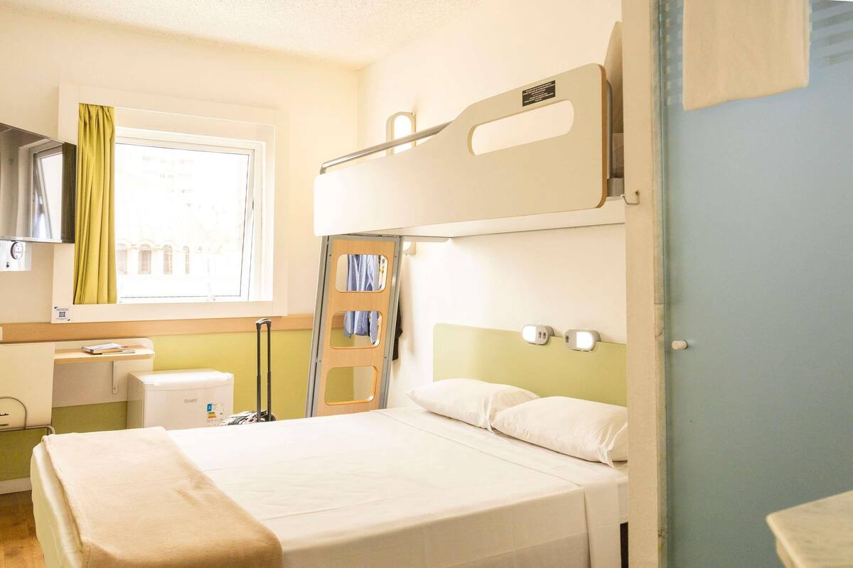 Une chambre d’hôtel avec un lit superposé, une fenêtre avec des rideaux, un bureau avec une chaise et une salle de bain.
