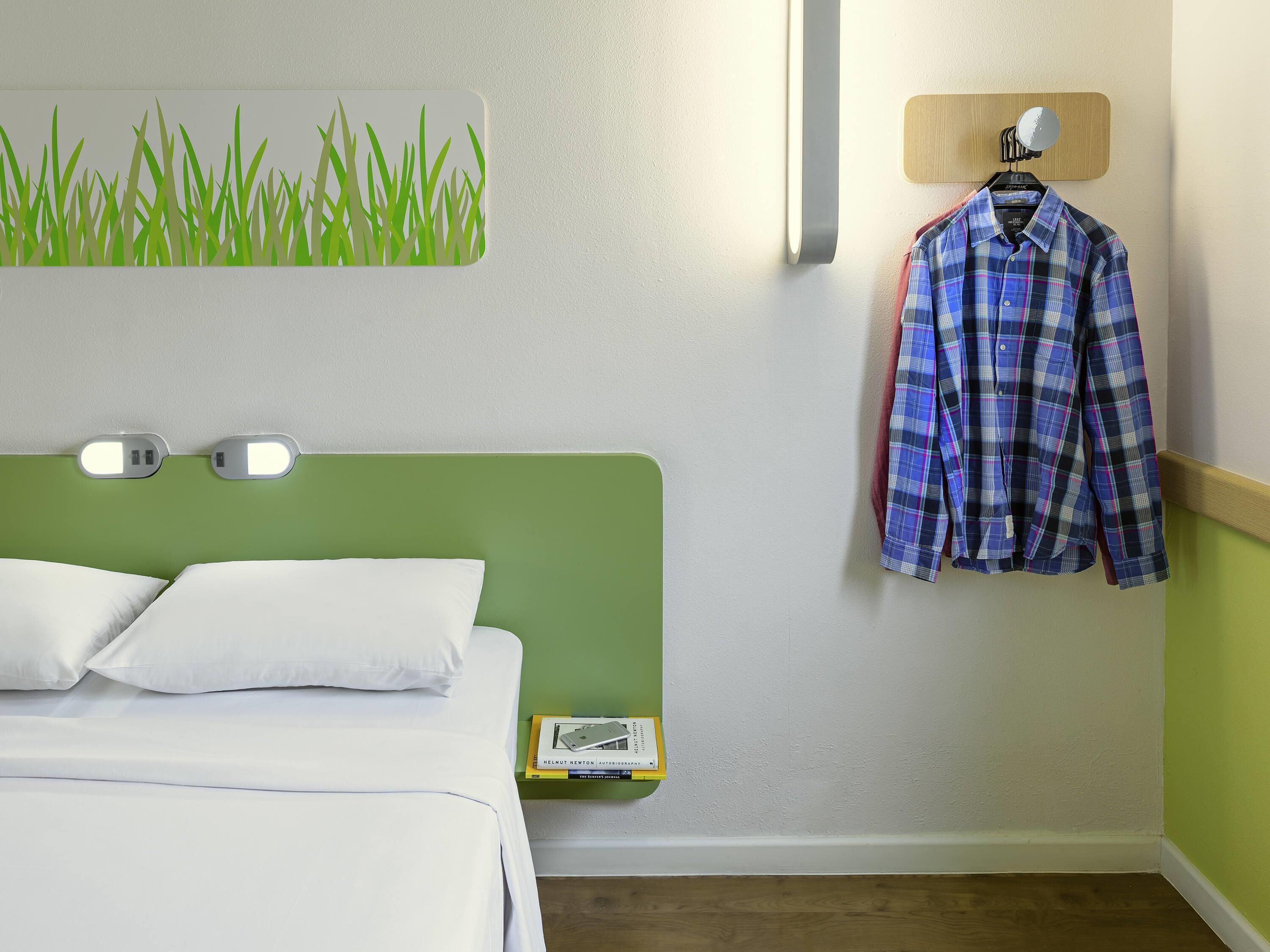 A hotel room with a green headboard, white bedding, a wall-mounted lamp, a hanging shirt, and a small bedside table with a book.