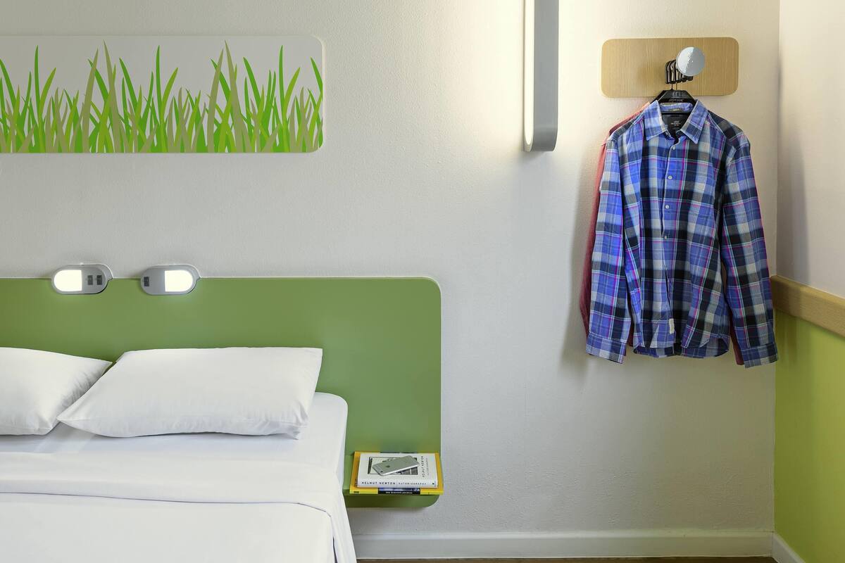 Hotelkamer met een groen hoofdeinde, witte beddengoed, een wandlamp, een hangend overhemd en een klein nachtkastje met een boek.