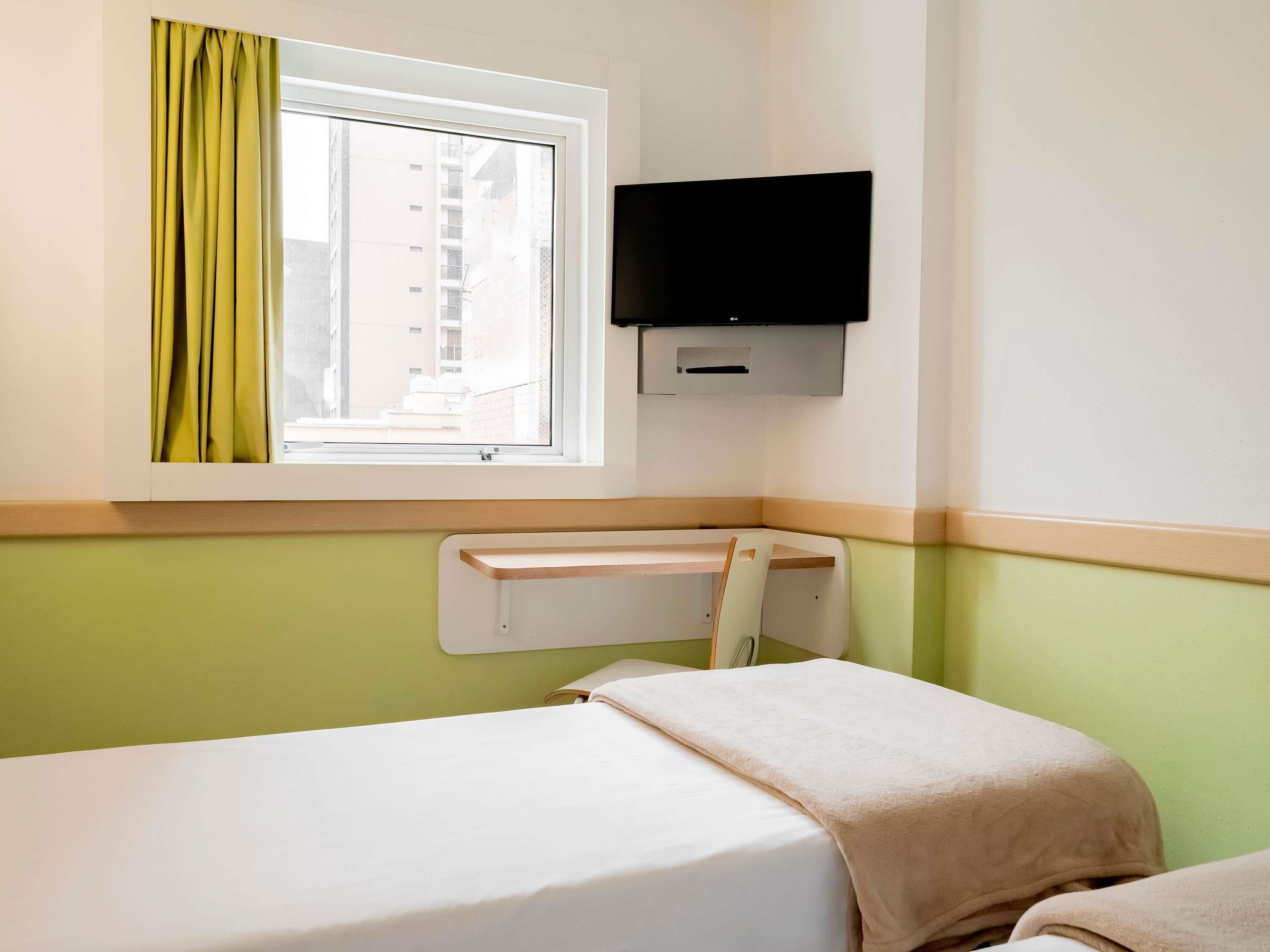 Ein Hotelzimmer mit einem Einzelbett, einem Fenster mit Vorhängen, einem Wand-Fernseher und einem kleinen Schreibtisch.