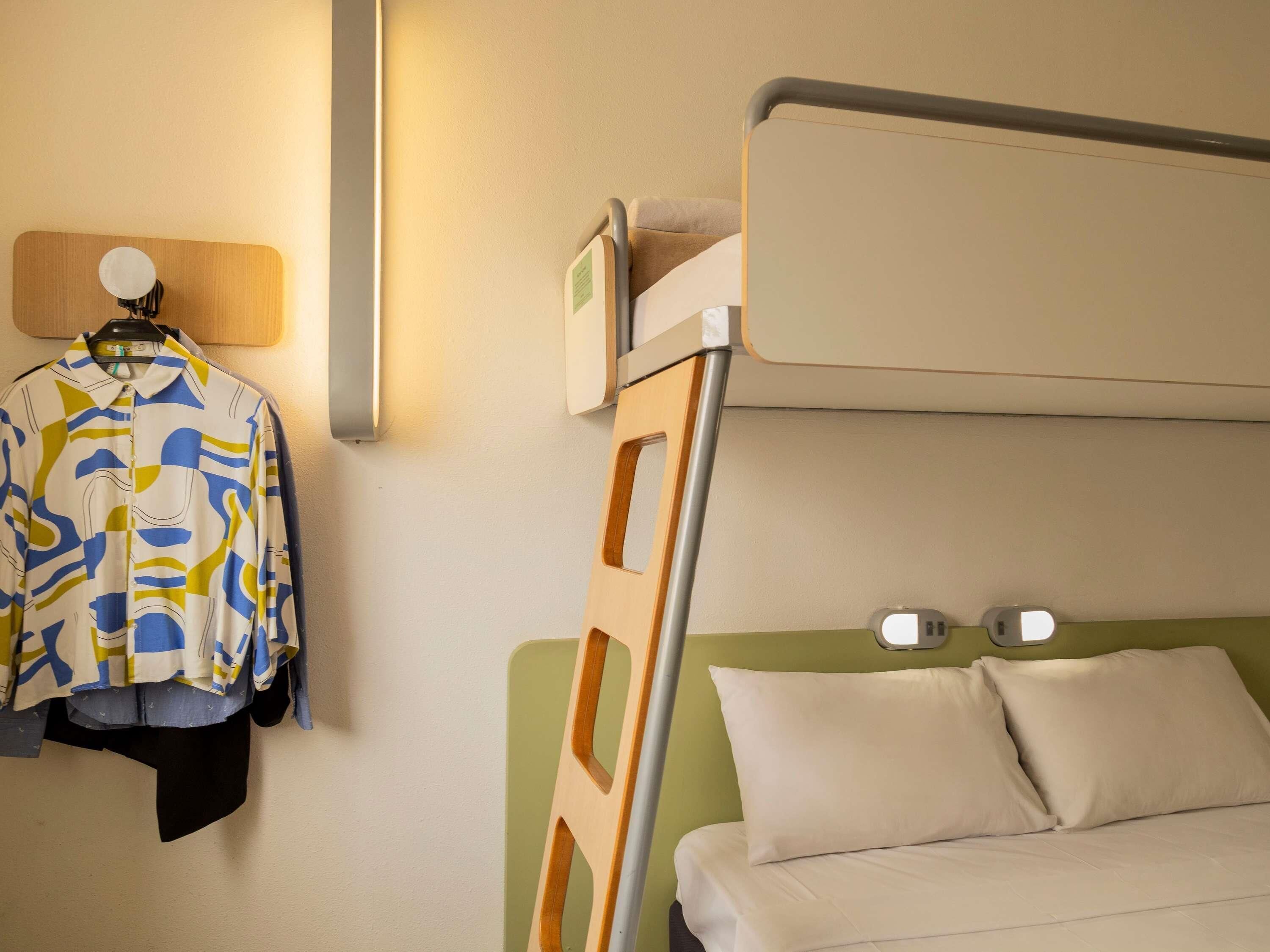 A hotel room with a bunk bed, a hanging coat rack, and a bedside table with a lamp.