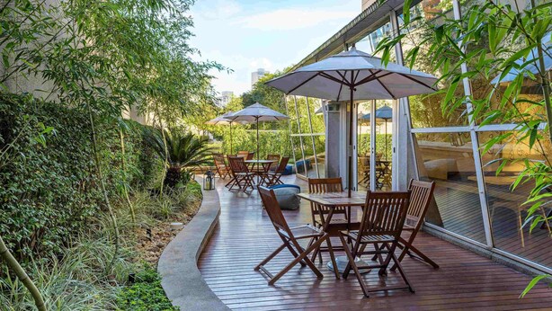 Een terras buiten met houten tafels en stoelen, omgeven door groen en een glazen balustrade.