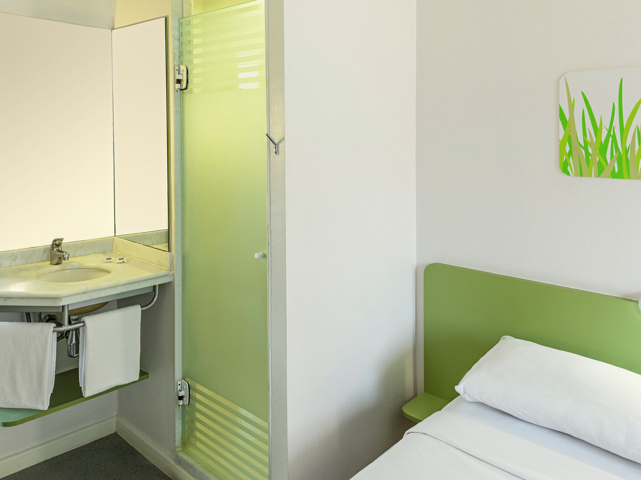 A hotel room with a bathroom including a sink, mirror, and shower. There is a bed with white bedding and a green headboard.
