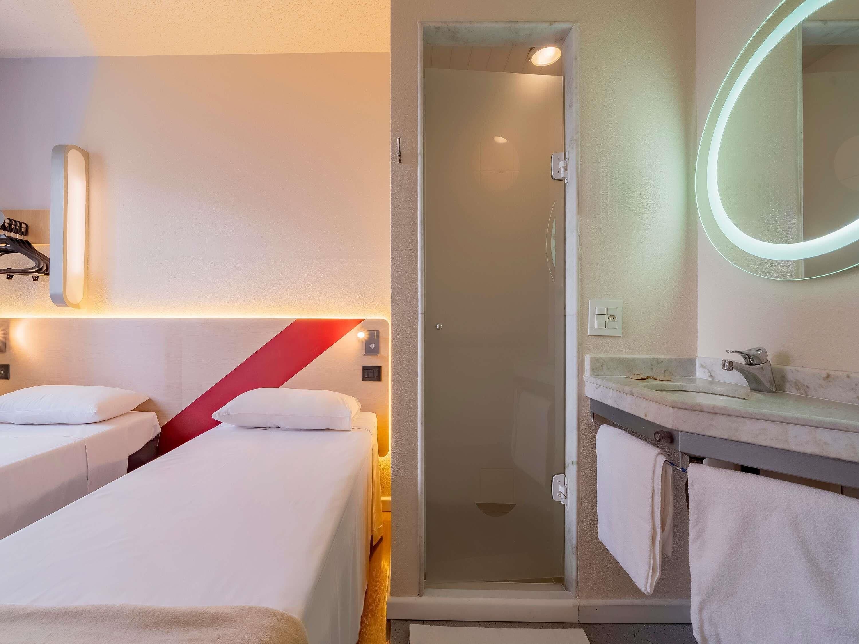 Habitación de hotel con dos camas, ducha, baño con lavamanos y espejo, y una pared de acento roja.