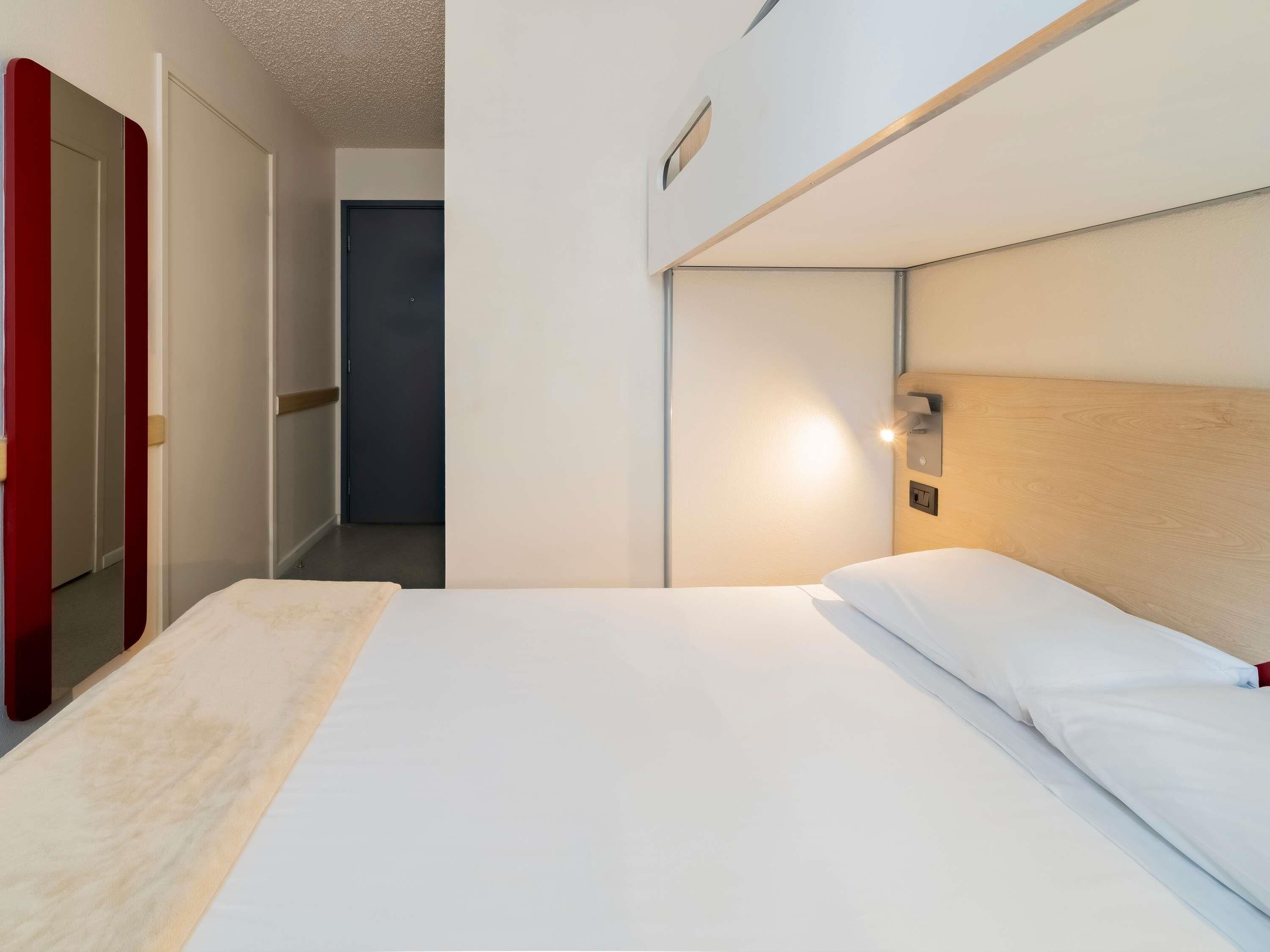 Habitación de hotel con una cama, cabecera de madera, mesita de noche y espejo con marco rojo.