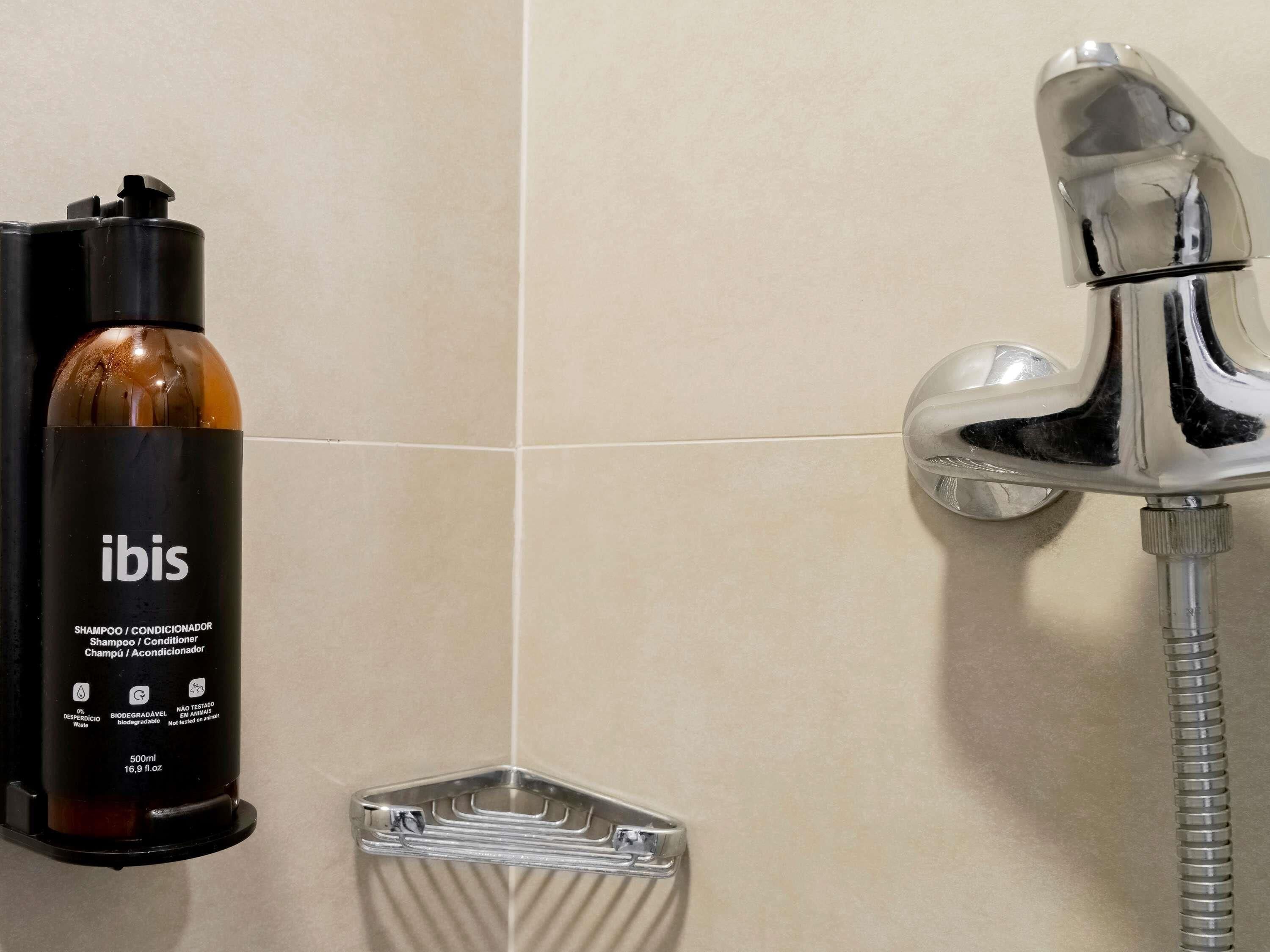 Une salle de bain avec un flacon de shampoing noir portant l’inscription « ibis », un robinet chromé et un receveur de douche transparent.