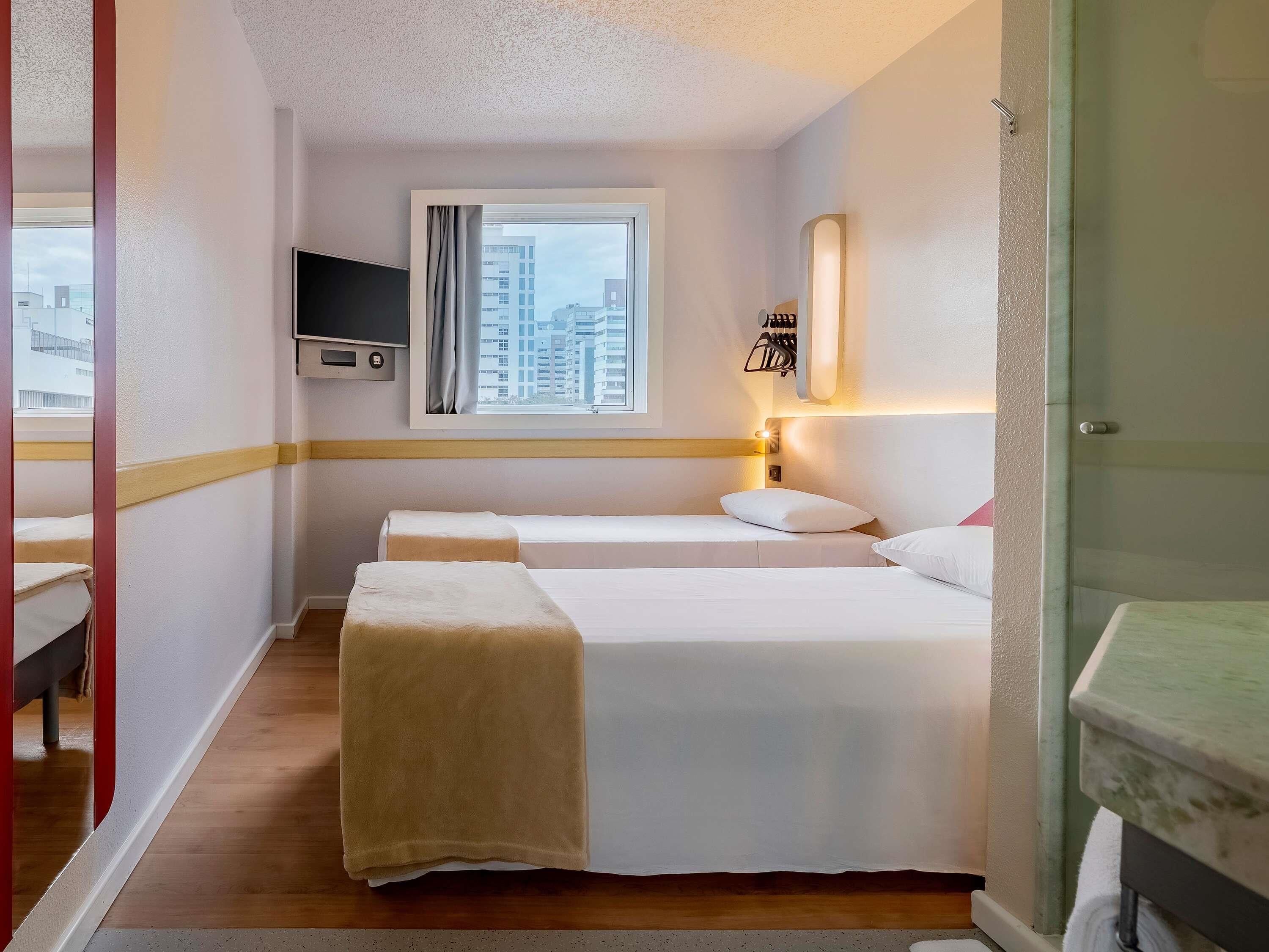 Habitación de hotel con dos camas, un televisor montado en la pared, una ventana con cortinas y un baño con lavamanos.