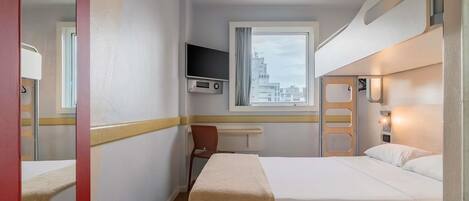 A hotel room with a bed, a TV mounted on the wall, a window with a view of buildings, and a chair.