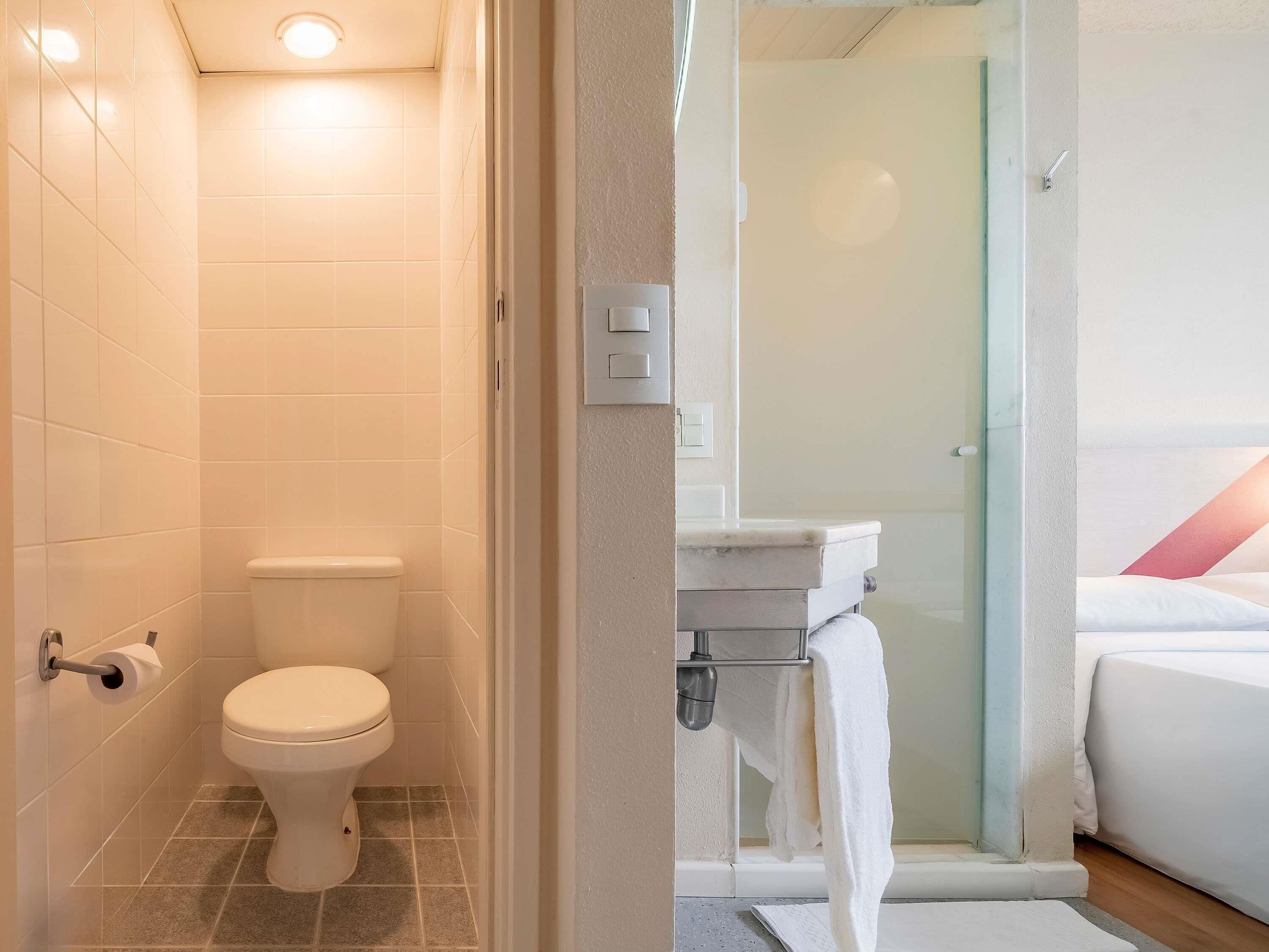 A bathroom with a toilet, a shower area, and a bed with a white mattress and a red pillow.