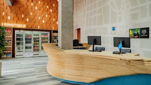 A modern reception area with a wooden reception desk, a large pillar, and a counter with a blue flag. There are refrigerated display cases in the background and a wall with a cityscape mural. - ibis budget Sao Paulo Paulista (São Paulo)