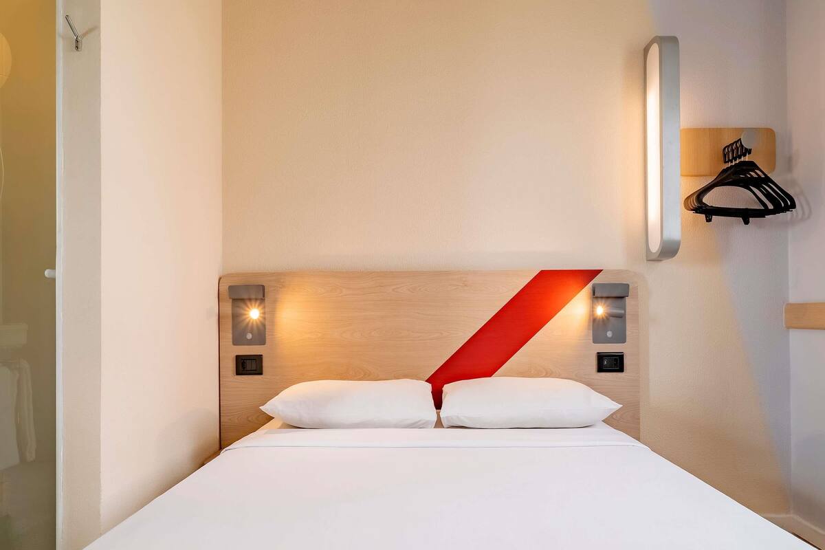 A hotel room with a bed, a wooden headboard, a red accent, wall-mounted lights, and a coat rack.