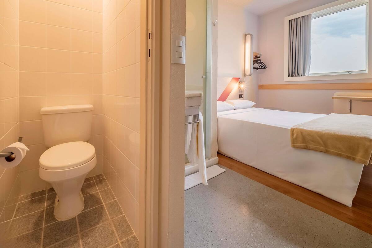 A bathroom with a toilet, a shower, and a bed with a window.