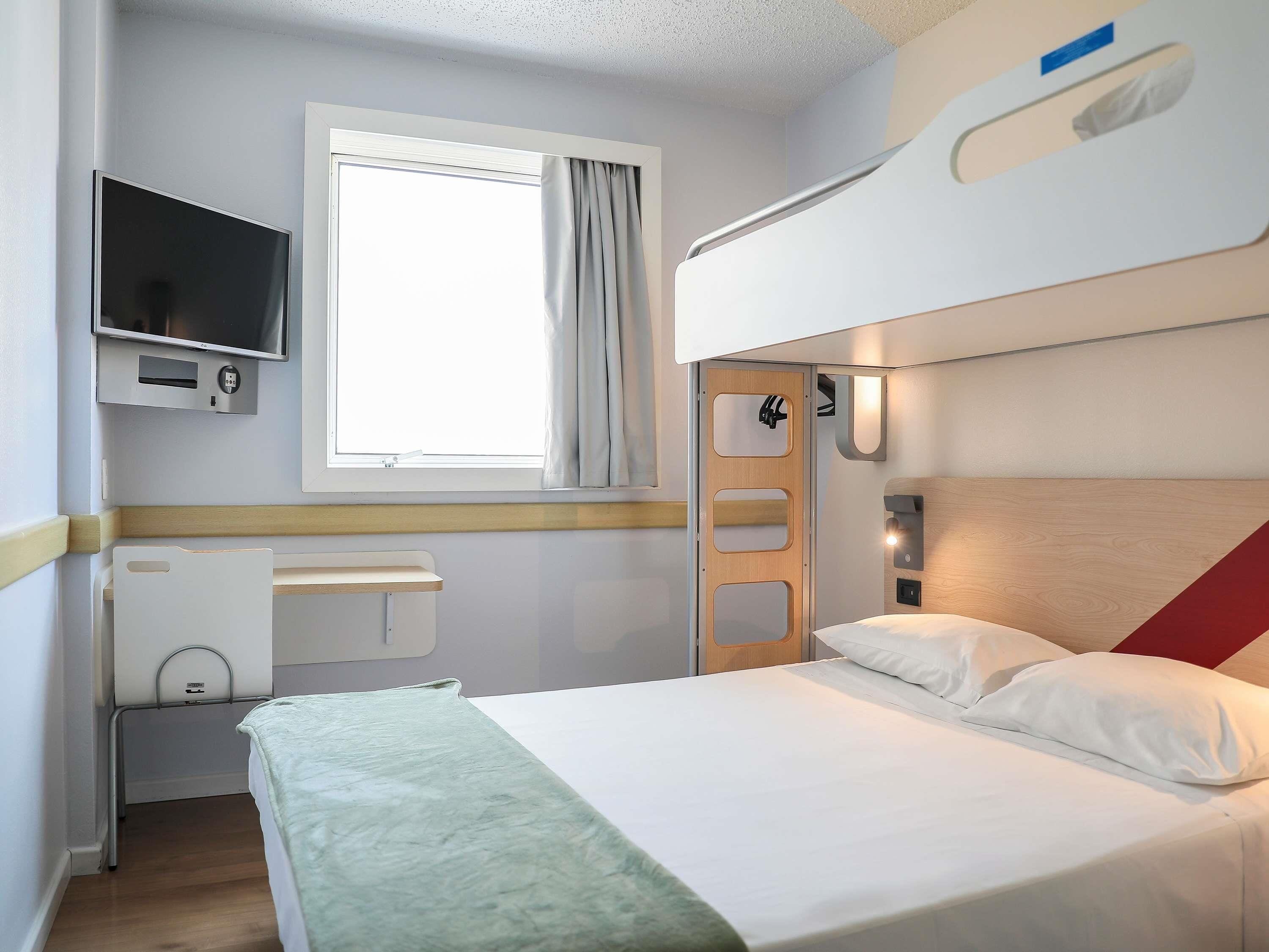 Uma cama beliche com uma televisão acima, uma janela com cortinas e uma mesa de cabeceira com tomada.