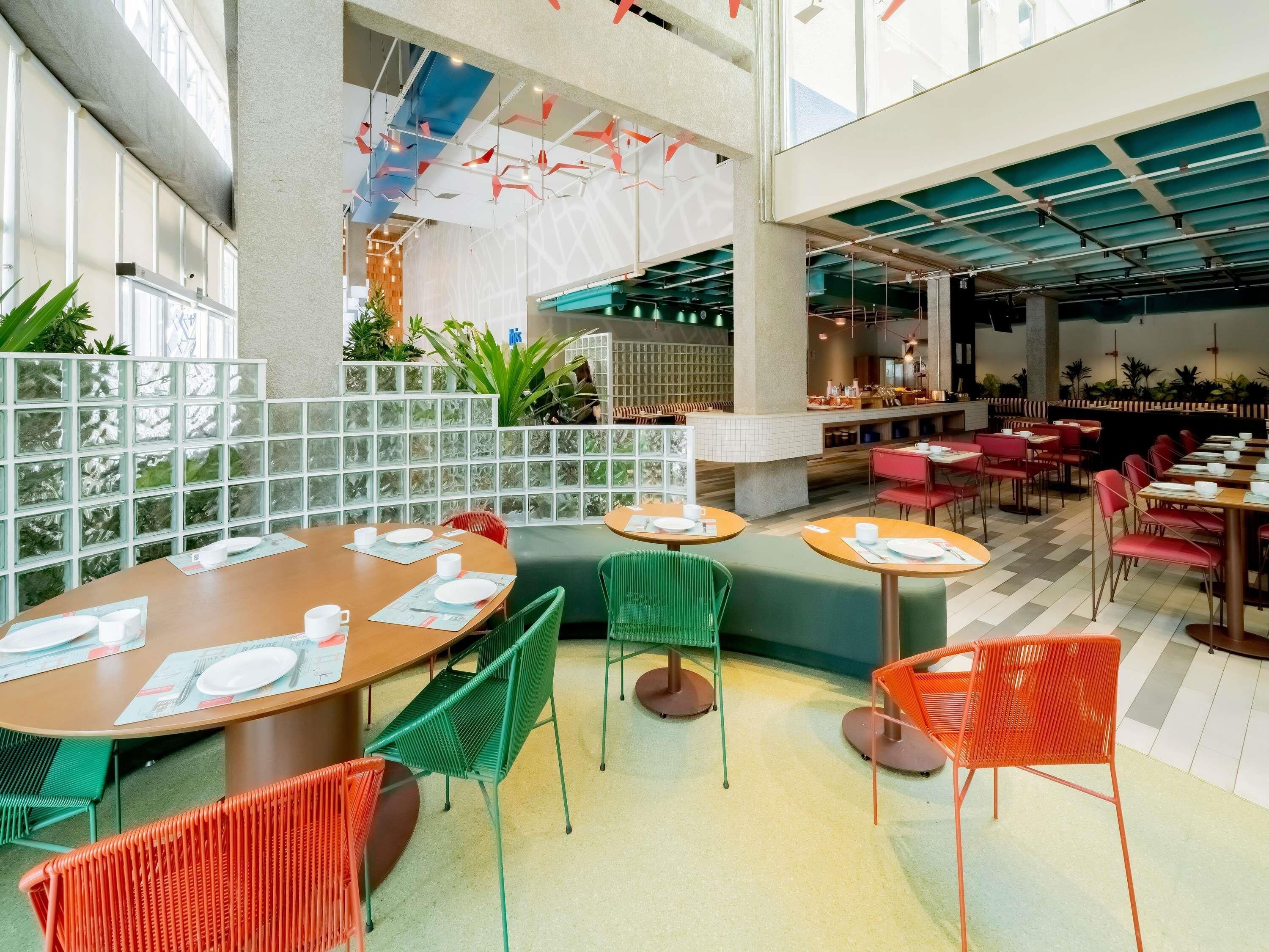 Un espace de restauration moderne avec des tables rondes, des chaises vertes et orange, et un plafond haut avec des poutres apparentes.