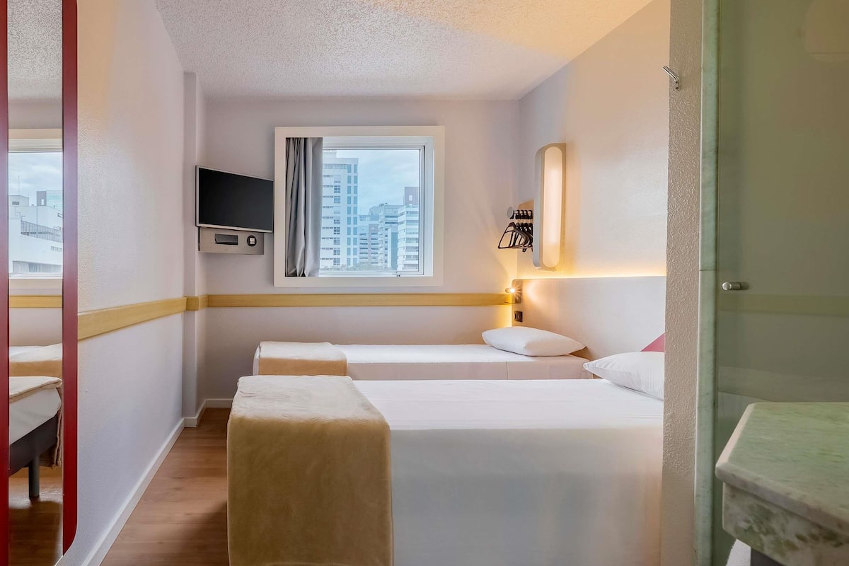 A hotel room with two beds, a TV mounted on the wall, a window with curtains, and a bathroom with a sink and mirror.