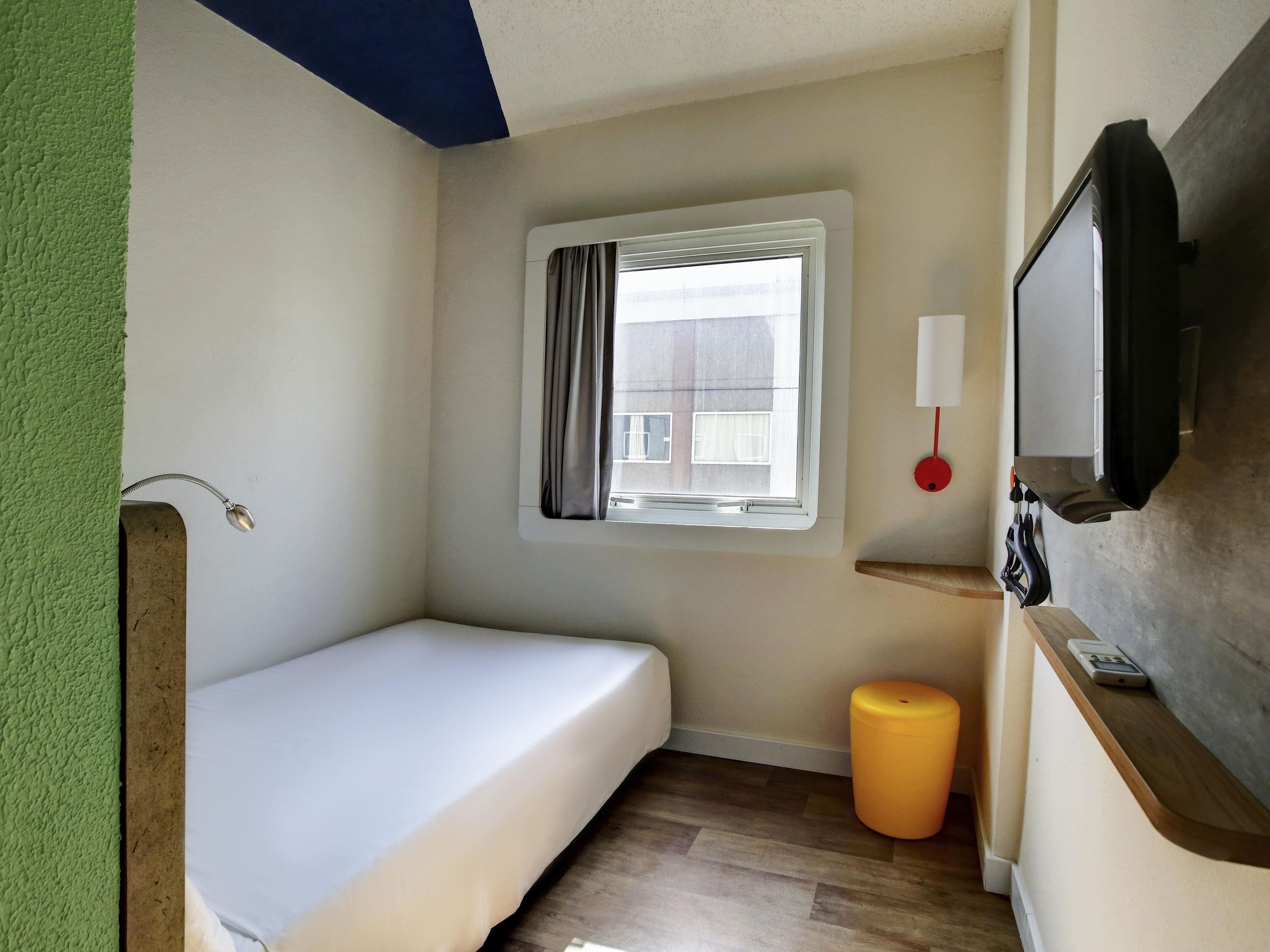 A small room with a single bed, a window with curtains, a wall-mounted TV, and a wooden shelf with a lamp.