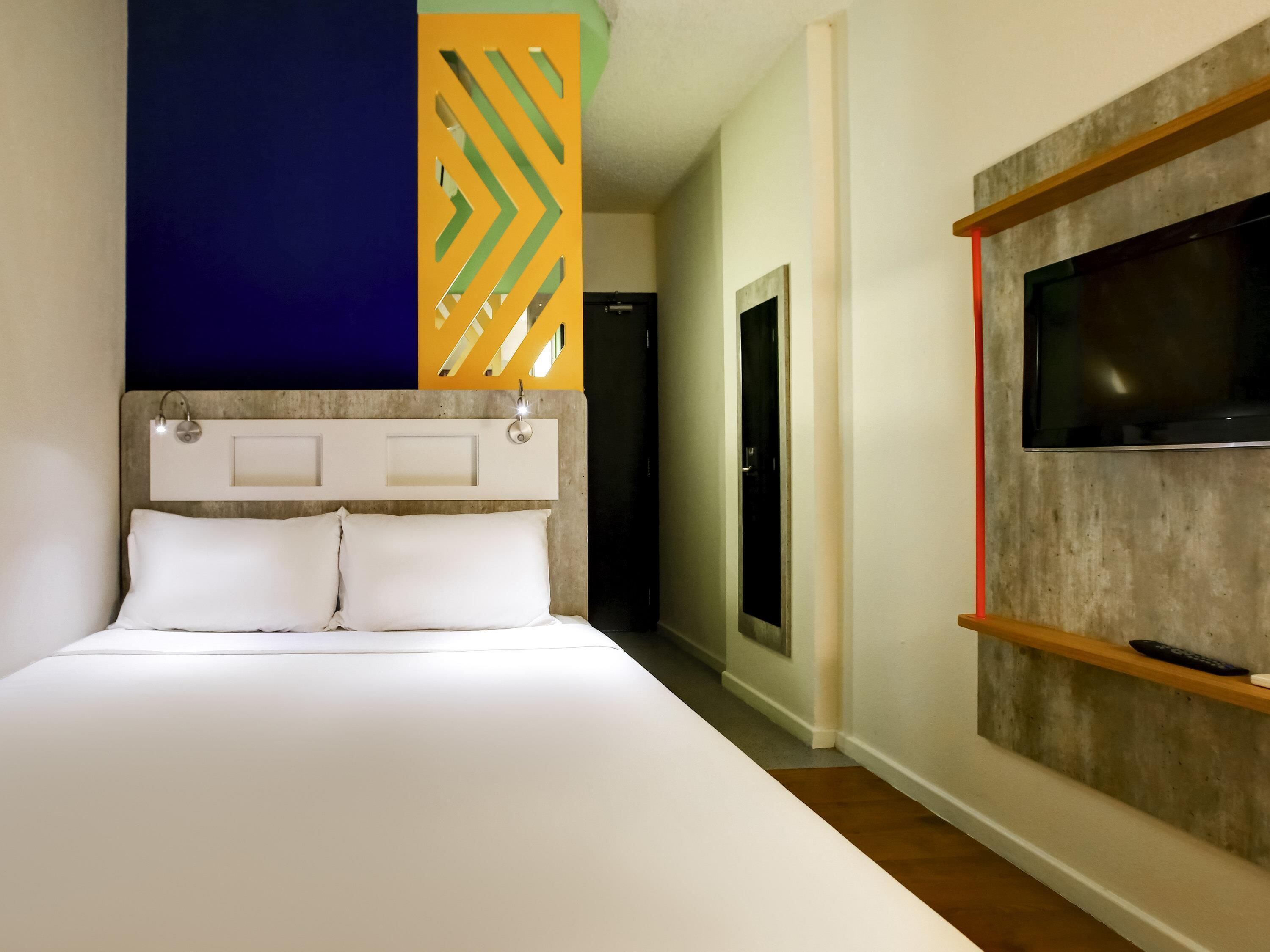 A hotel room with a bed, a TV mounted on a wall, and a hallway leading to a door.