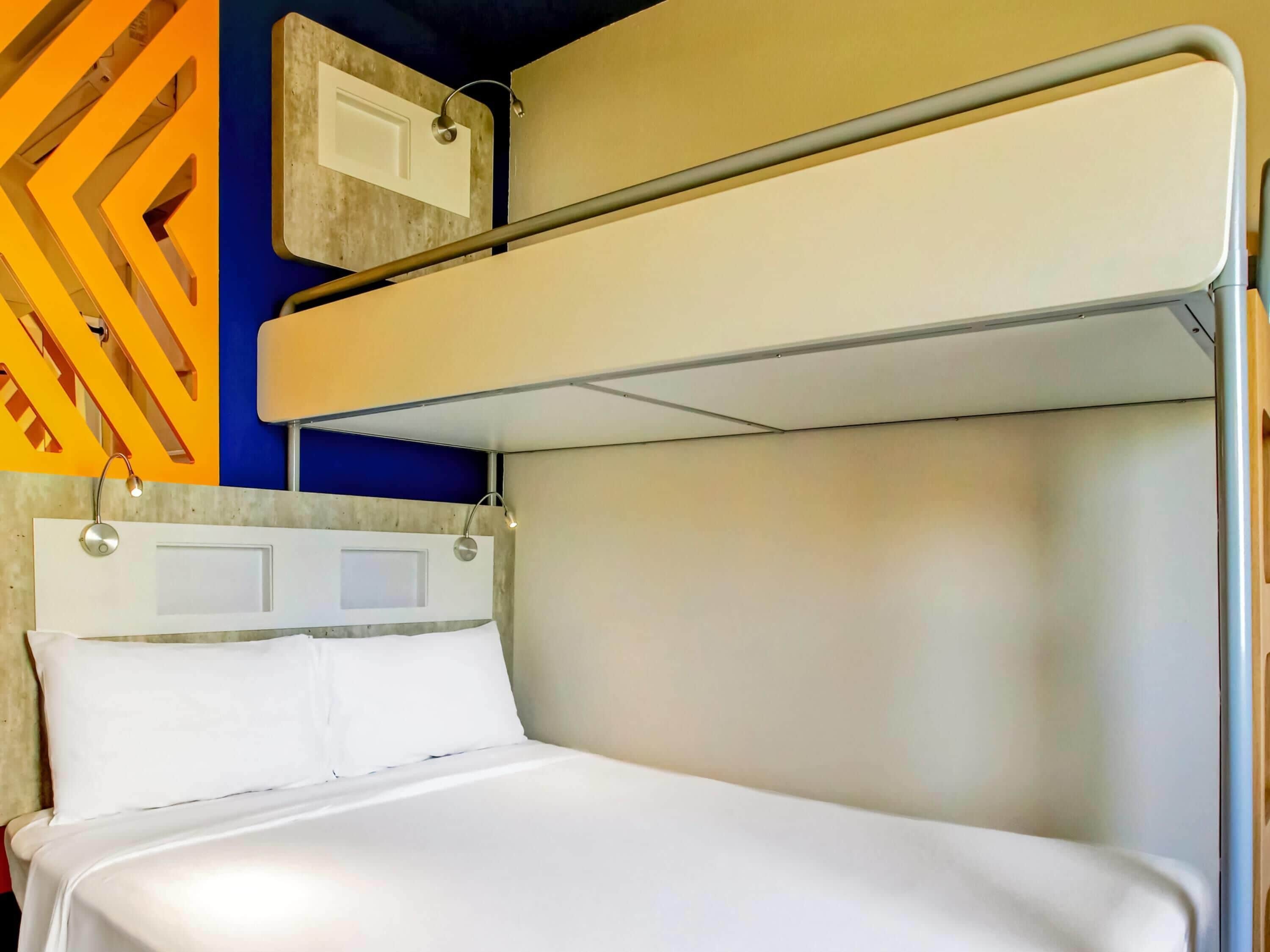 Una cama litera con colchón y almohadas blancas, un cabecero con una lámpara y una pared con diseño geométrico.