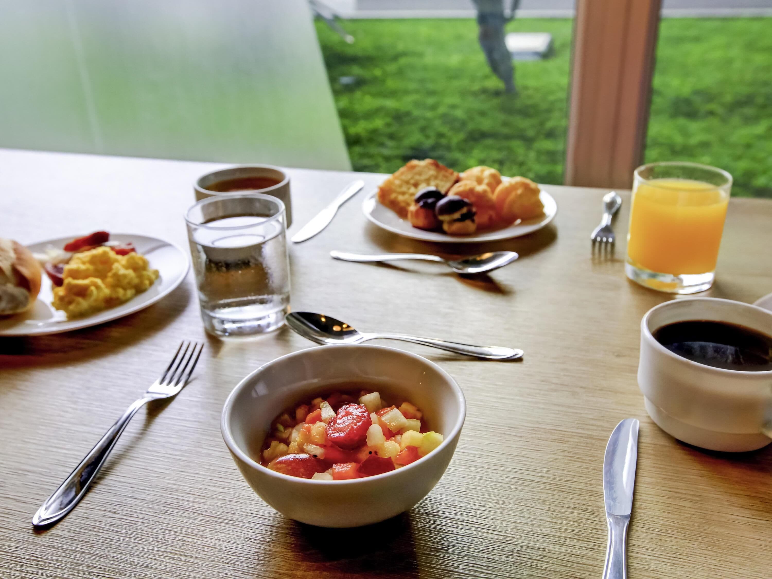 A breakfast table with plates of food, a bowl of fruit, a glass of orange juice, and cups of coffee and tea.