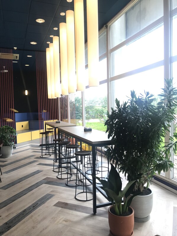 Un espacio interior moderno con una mesa larga y varios taburetes, ventanas amplias y plantas en macetas.