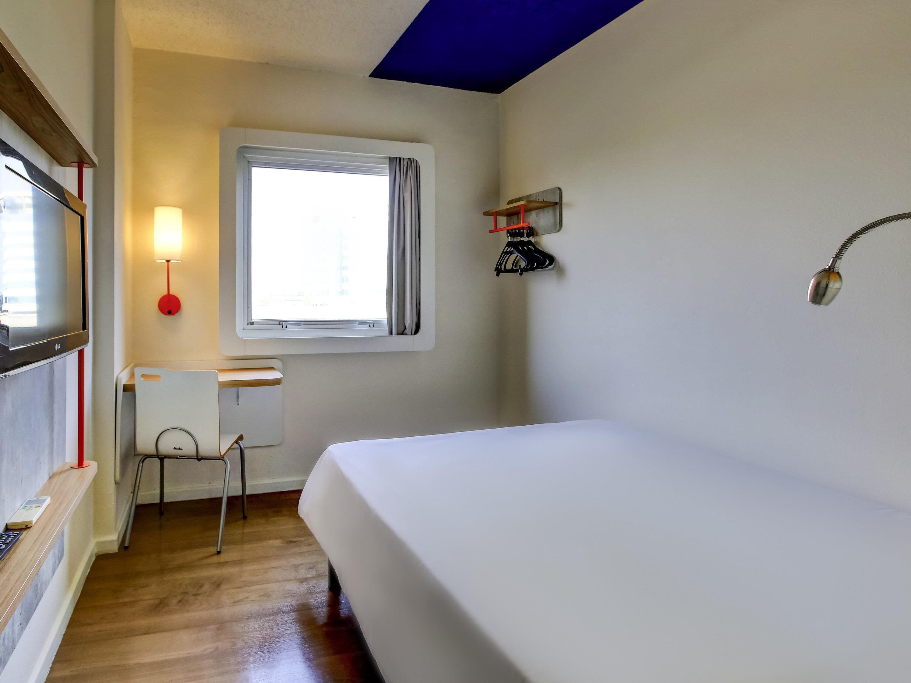 A bedroom with a bed, a desk, a chair, a window with curtains, a wall-mounted lamp, and a TV.